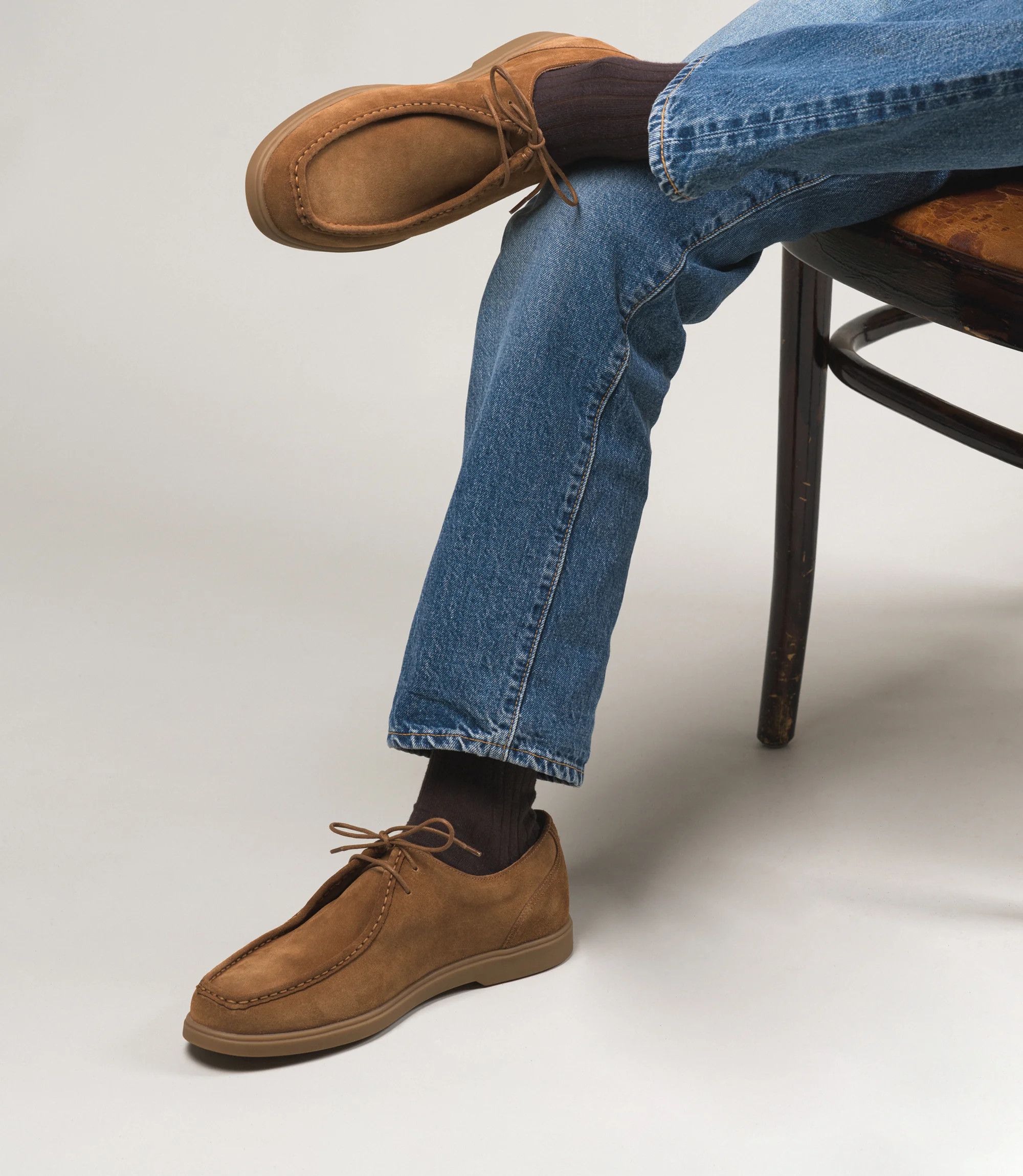 A man wearing light blue denim jeans sat down on chair in a studio with a grey backdrop wearing his Loake Arezzo shoes in Chestnut Suede.