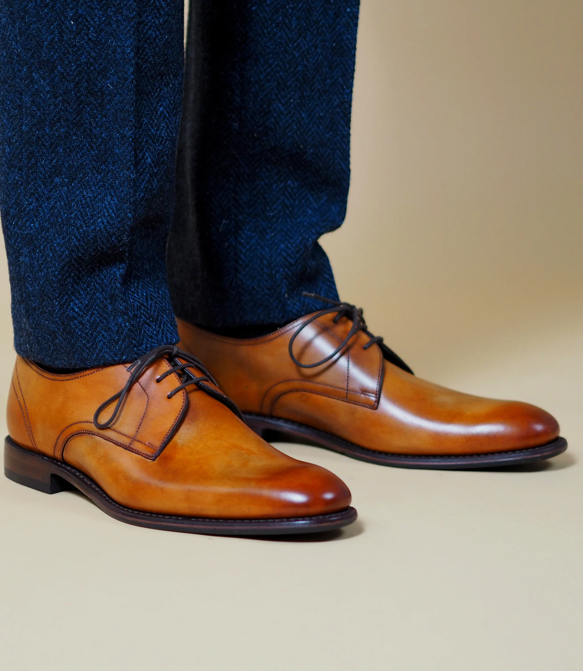 A man standing in a studio backdrop wearing navy trousers wearing his Loake Atherton in Tan leather.