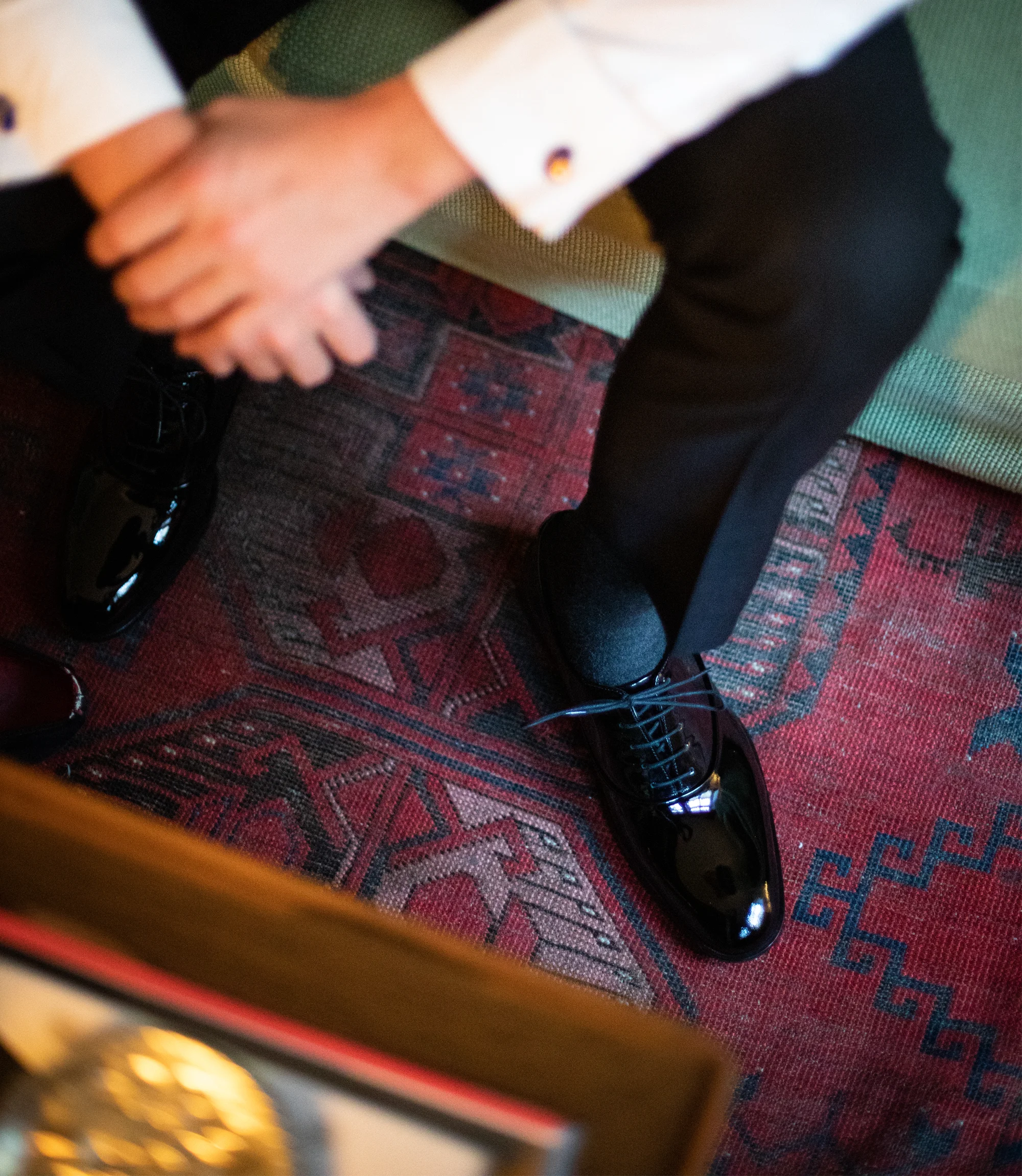 A man sitting down wearing black trousers and a white shirt in a living room wearing his Loake Patent shoes in Black Polished Leather.