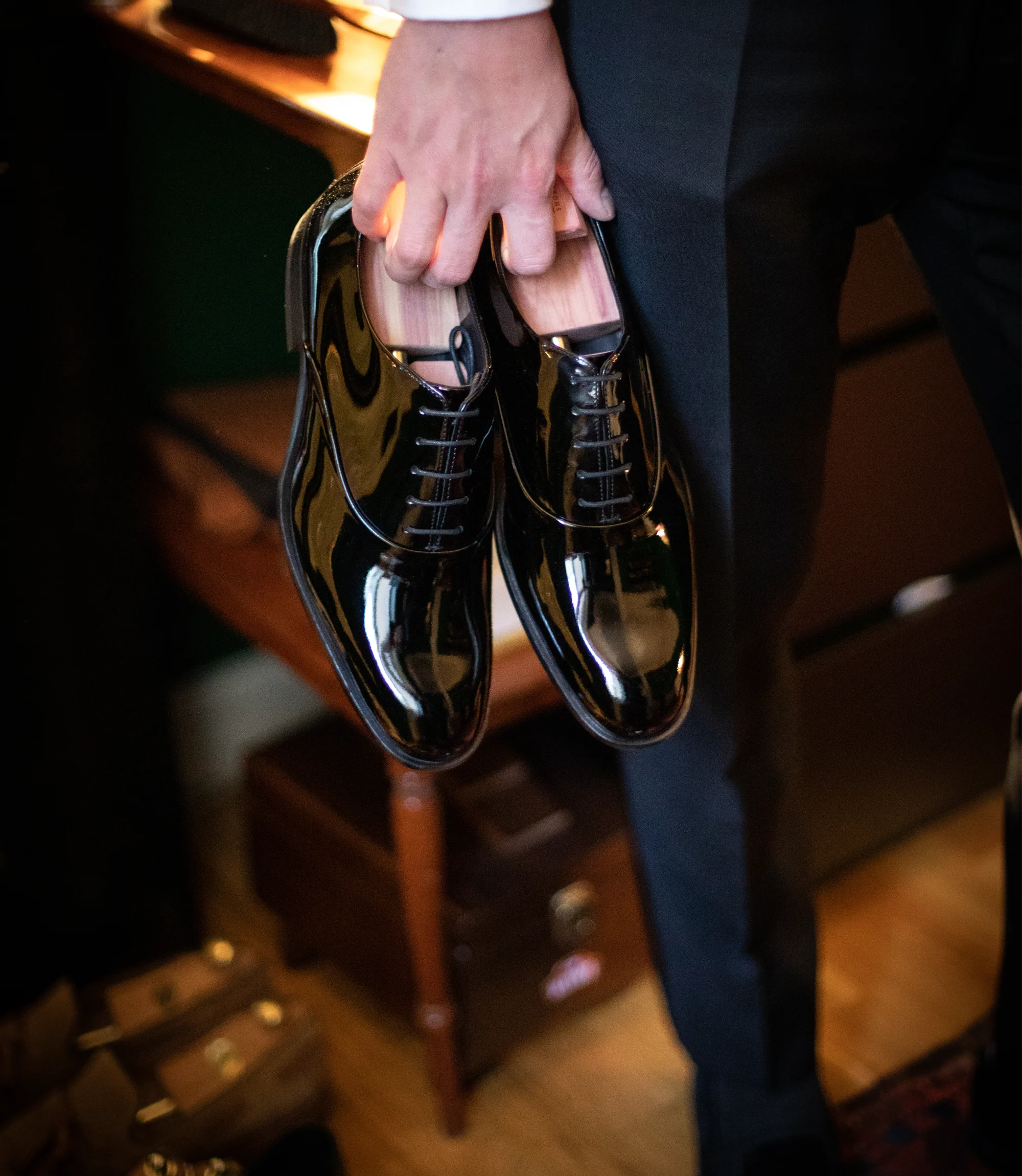 A man wearing black trousers holding a pair of Loake Patents in Black polished Leather with Loake shoe trees inserted.
