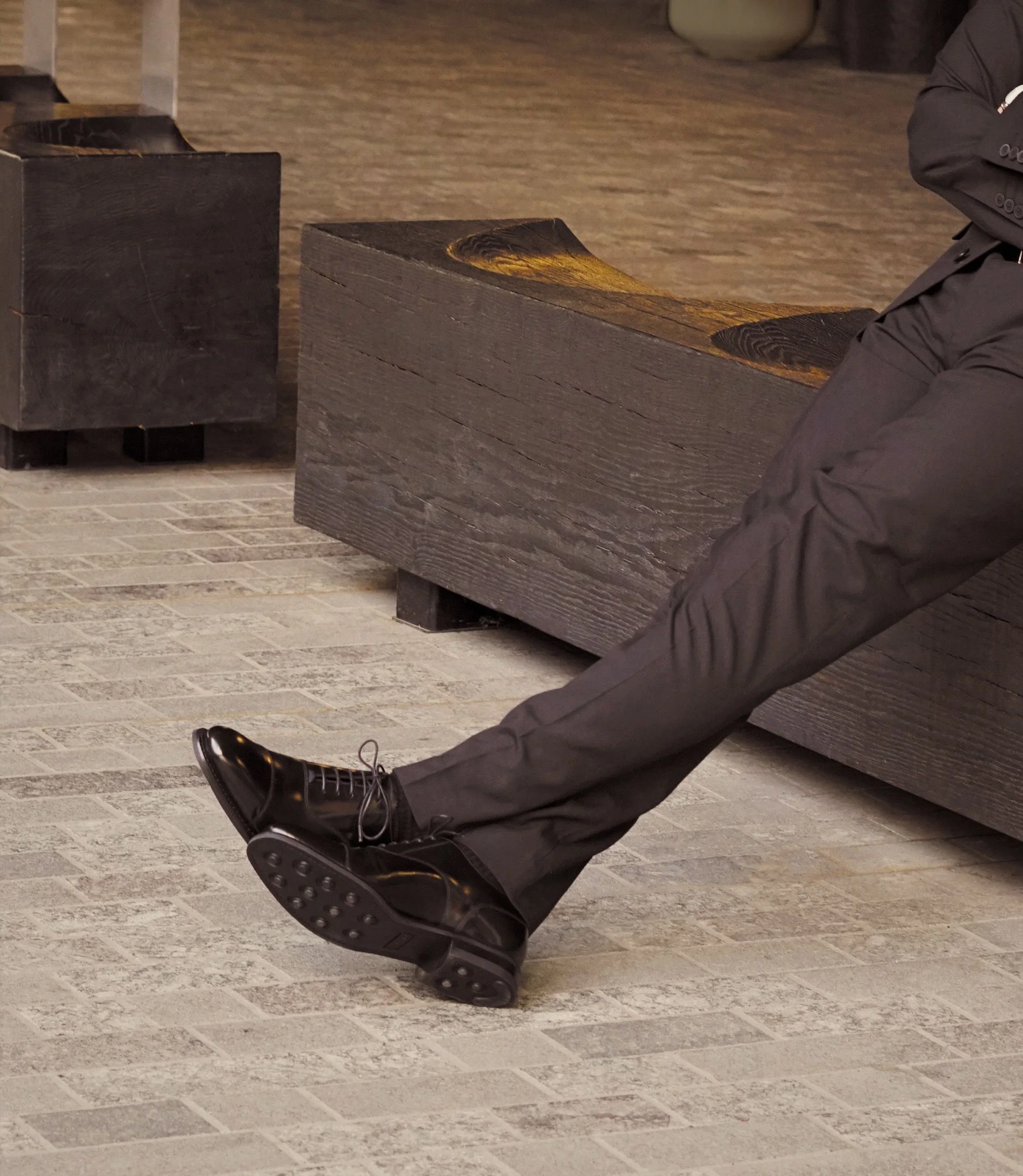 A man wearing black trousers sitting on a wooden bench outside in his Loake 300s in Black Leather.
