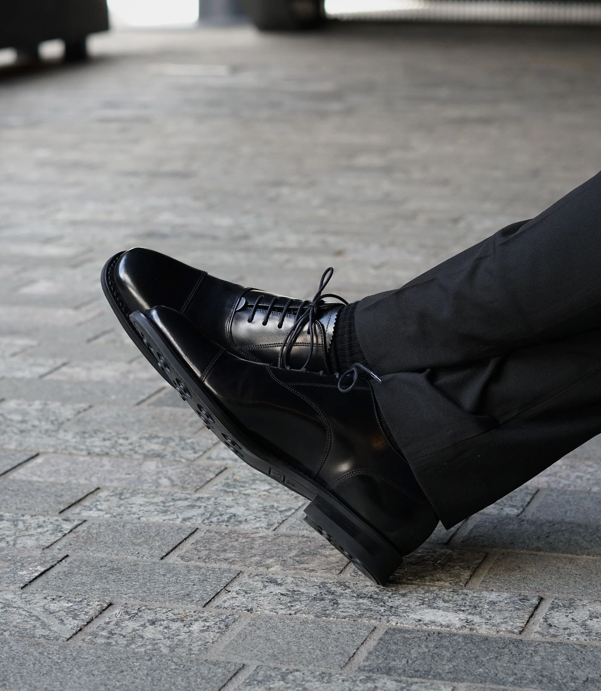 A man wearing black trousers sitting on a wooden bench outside in his Loake 300s in Black Leather.