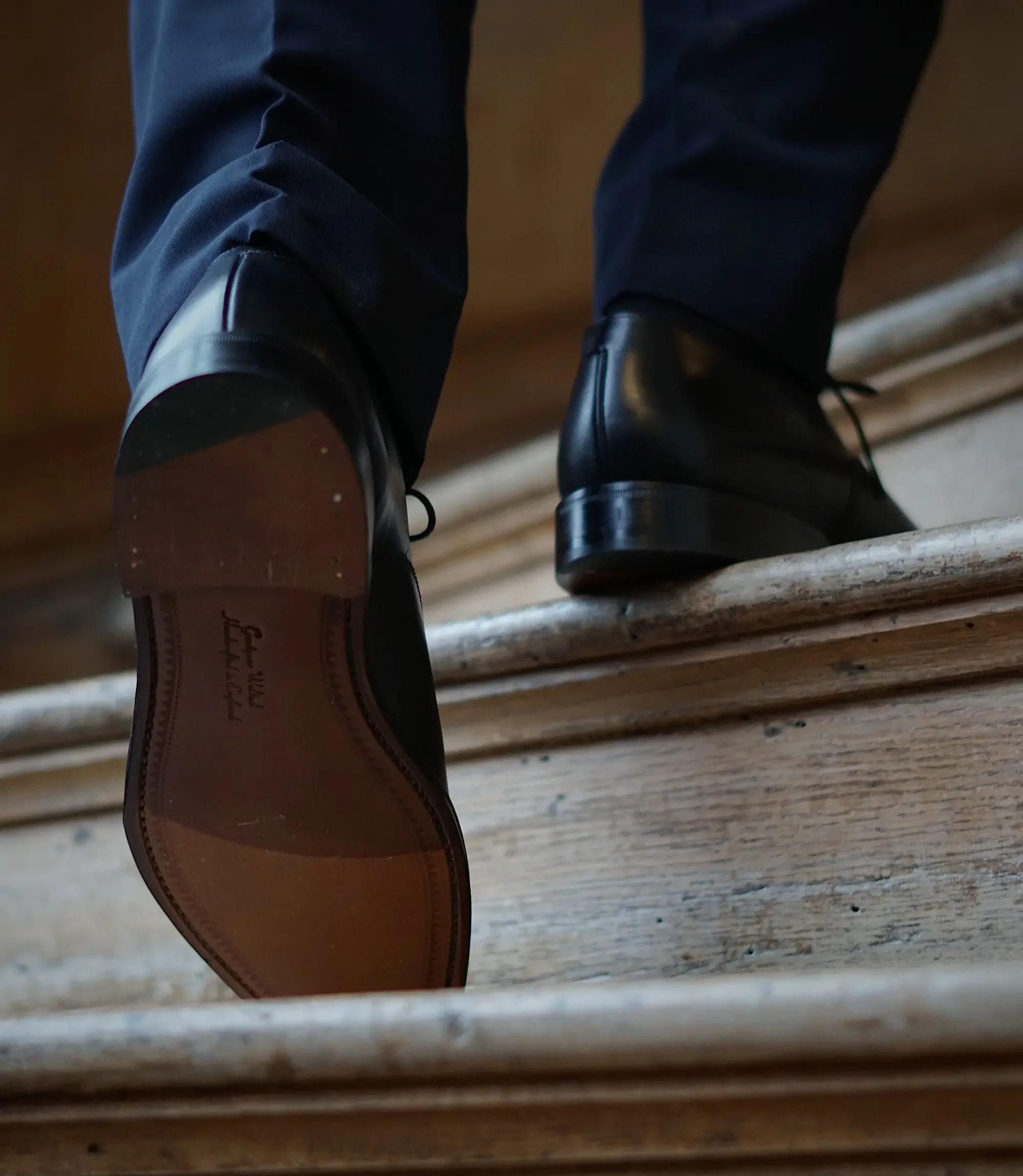 A man wearing navy trousers walking up wooden stairs wearing his Loake Aldwych black leather.