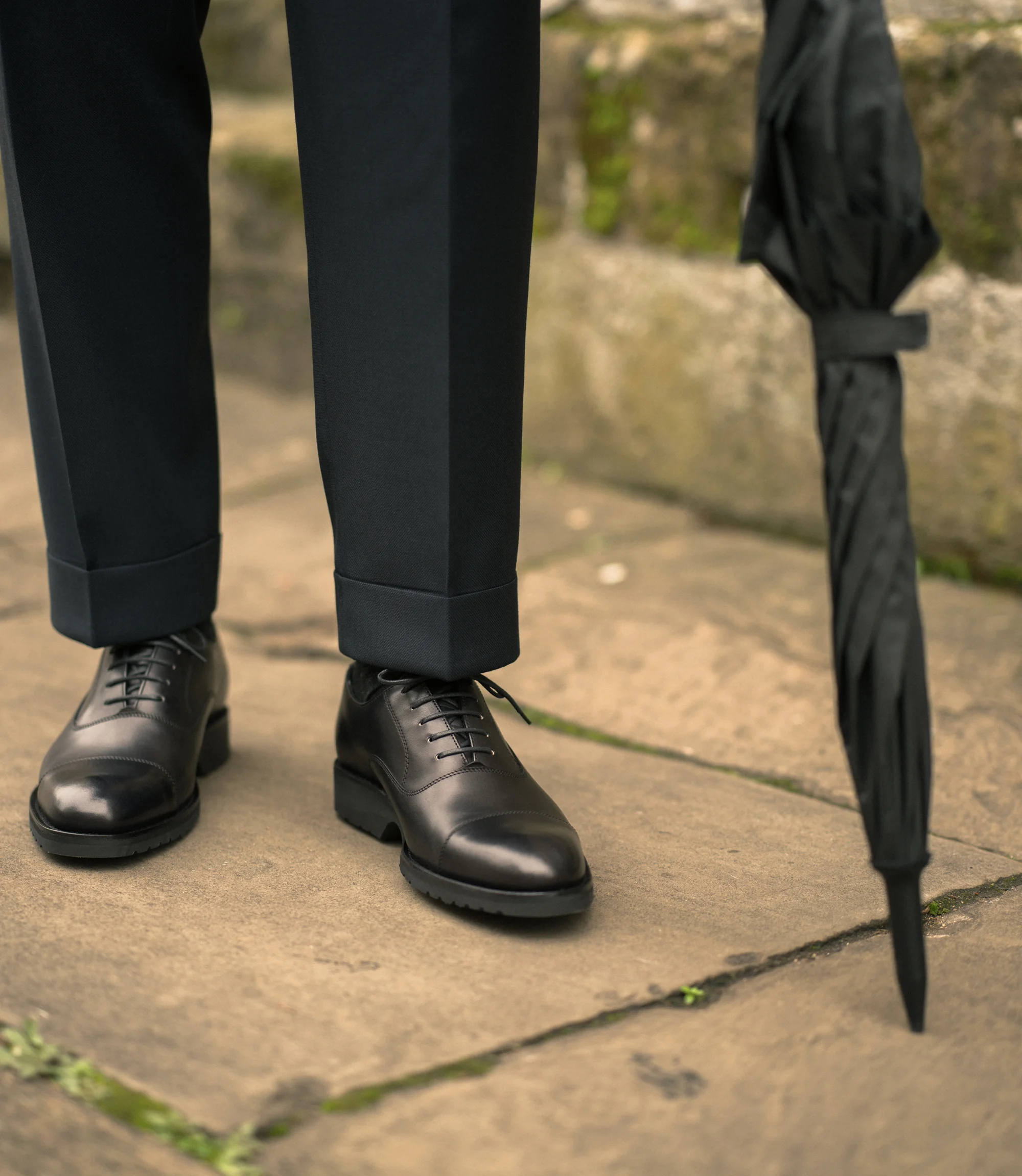 A man wearing navy trousers with an umbrella standing on concrete floor outside wearing his Loake Hitchcock in Black Leather.