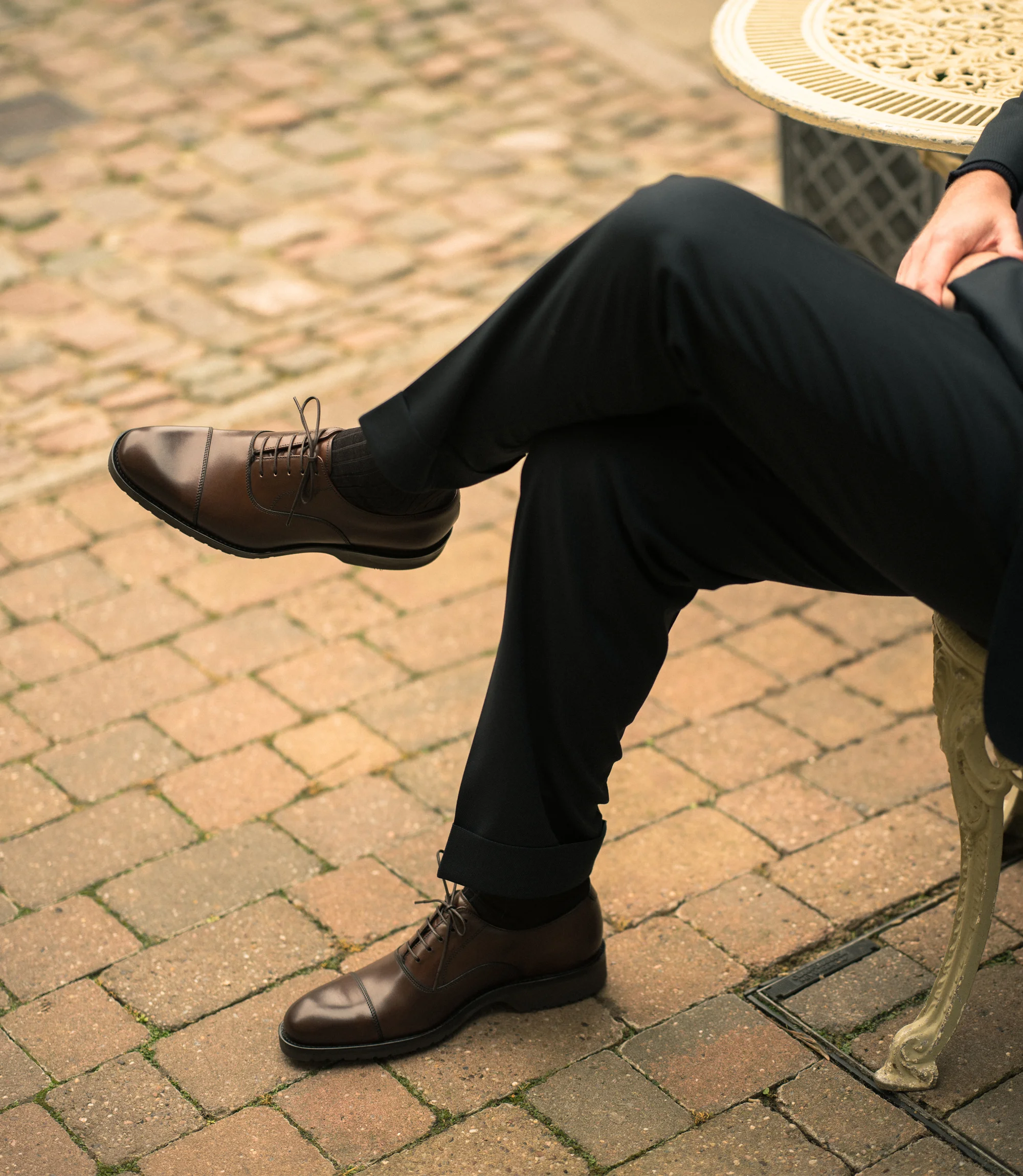 A man wearing black trousers sitting down on a chair outside wearing his Loake Hitchcock in Dark Brown Leather.