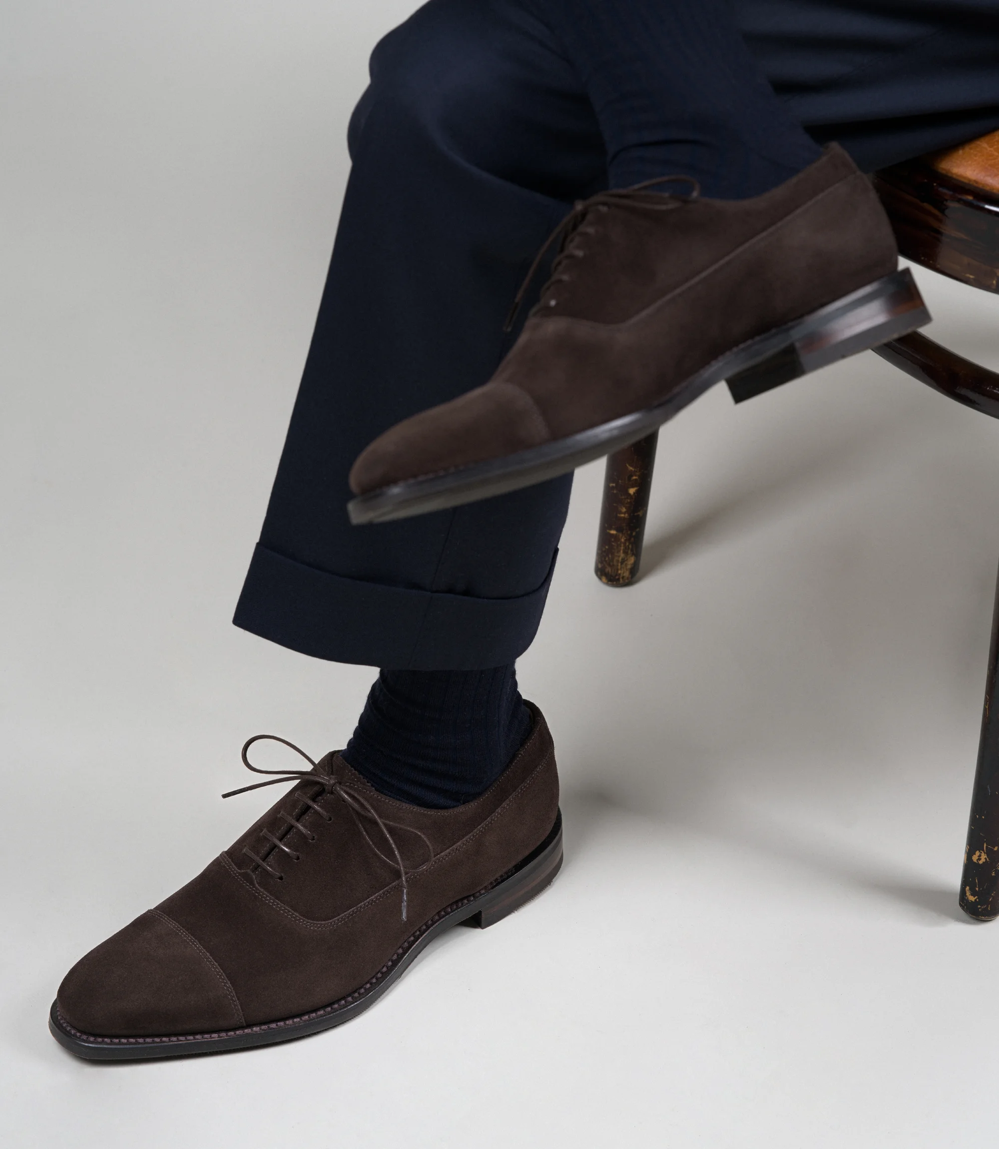 A man wearing navy trousers sitting on a chair in a studio with a grey backdrop wearing his Loake Truman Toecap shoes in Dark Brown Suede.