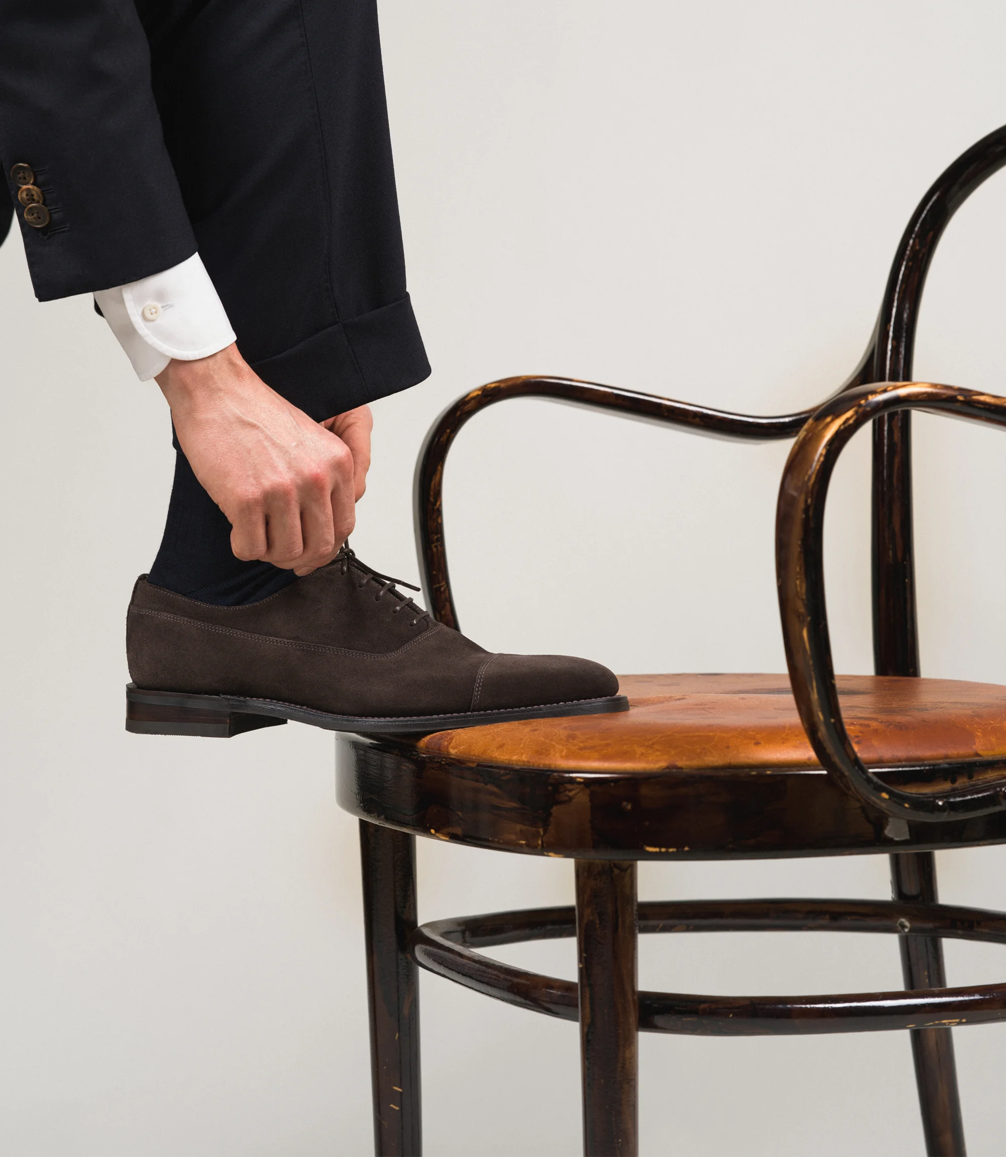 A man wearing navy trousers with his foot on a chair tying laces wearing his Loake Truman Toecap shoes in Dark Brown Suede.