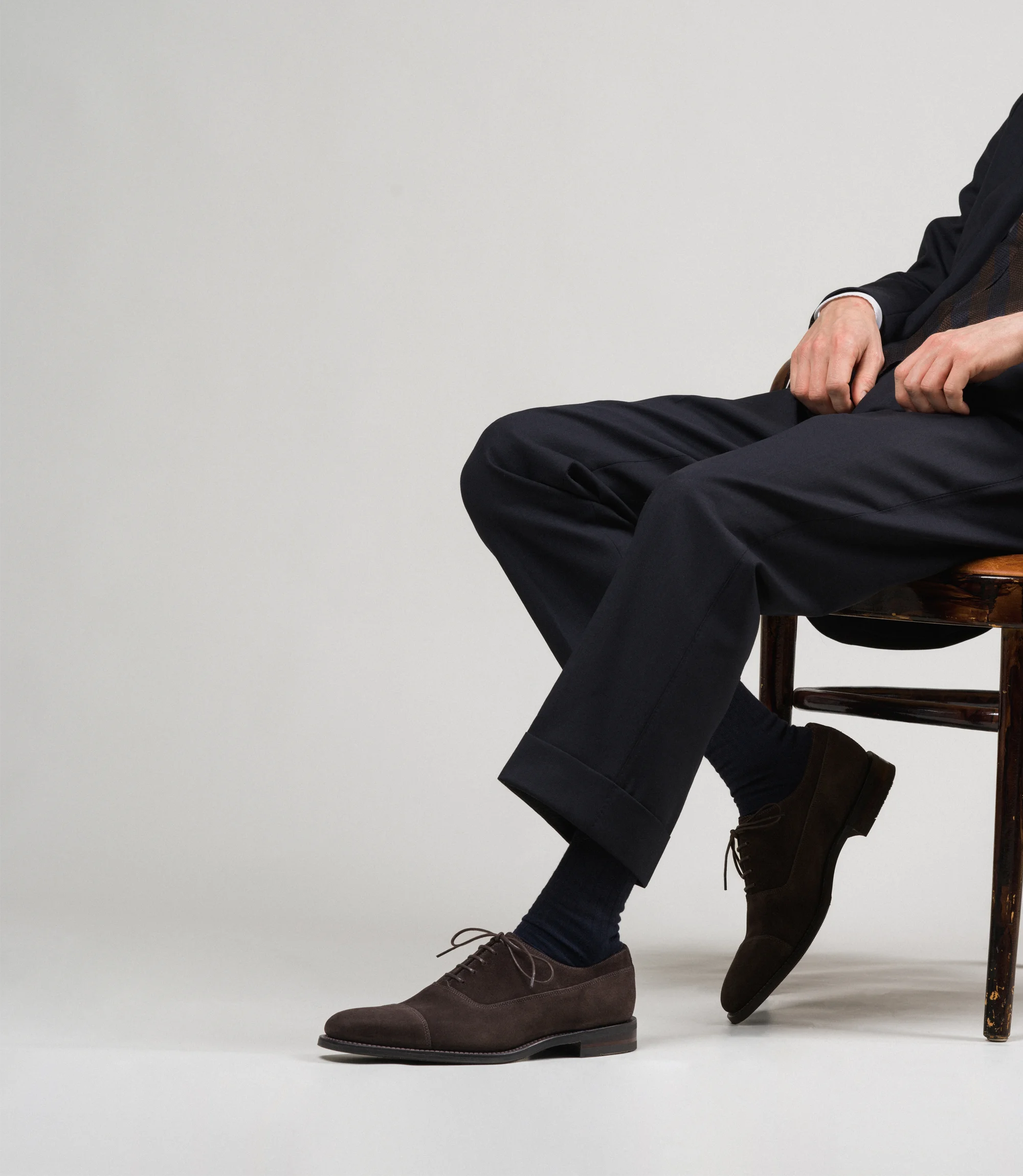 A man wearing navy trousers sitting on a chair in a studio with a grey backdrop wearing his Loake Truman Toecap shoes in Dark Brown Suede.