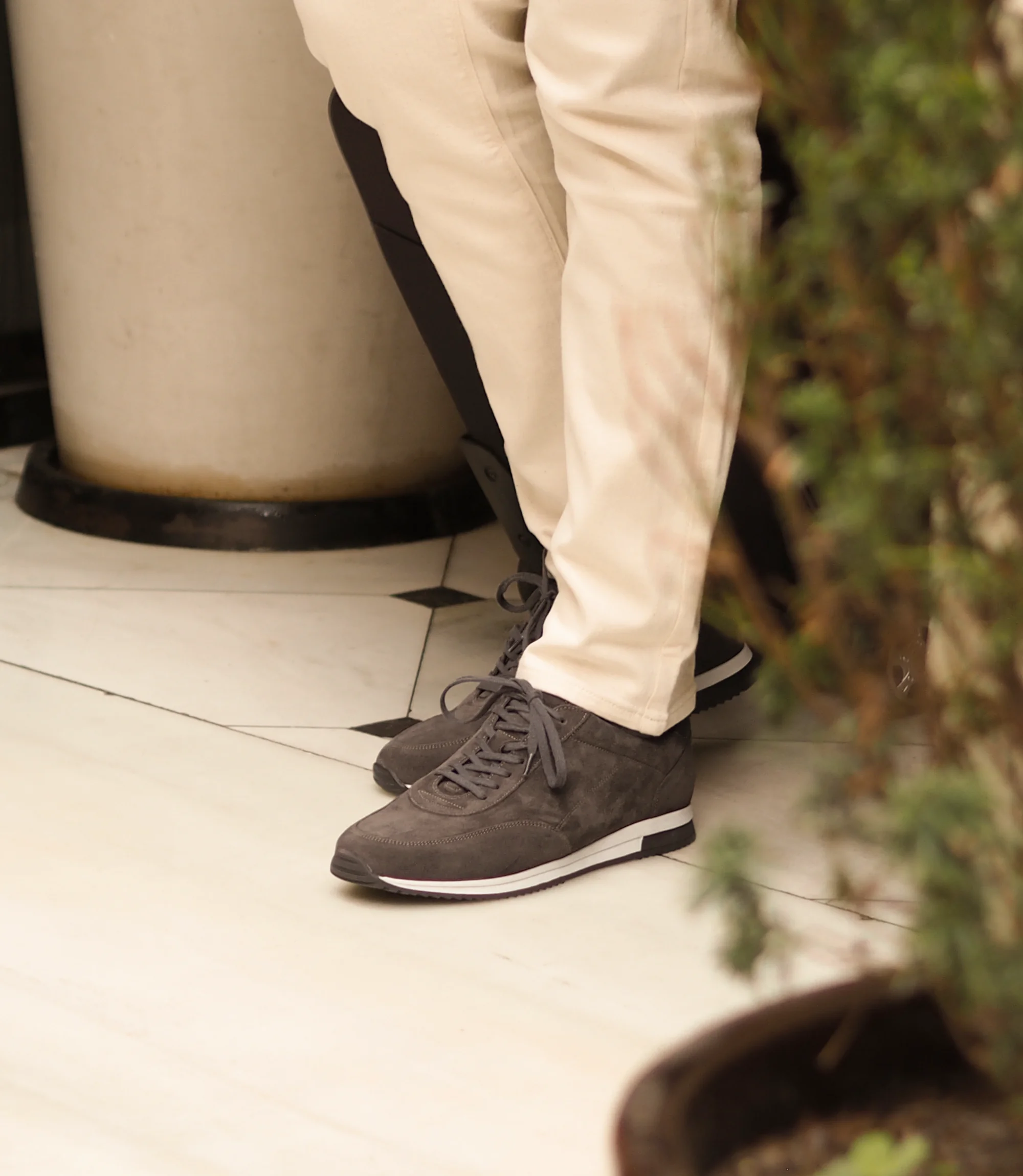 A man wearing white denim trousers standing with a suitcase on a marble floor wearing his Loake Bannister Trainers in Anthracite Suede.