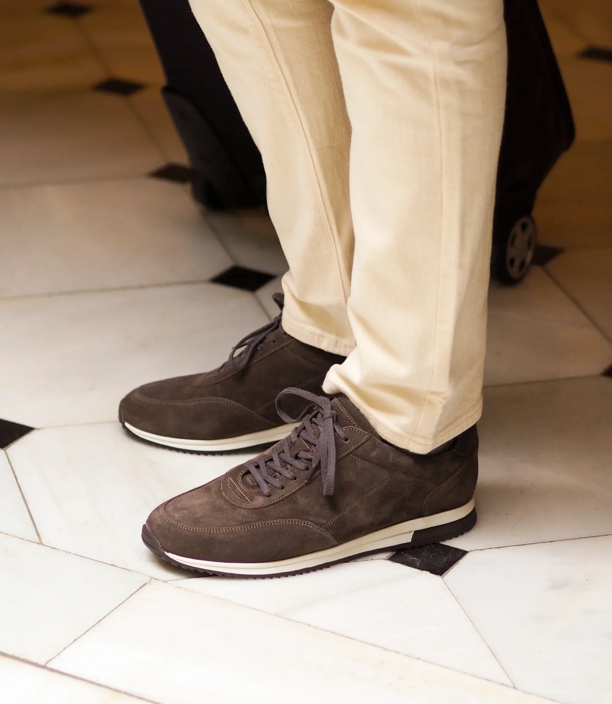 A man wearing white denim trousers standing with a suitcase on a marble floor wearing his Loake Bannister Trainers in Anthracite Suede.