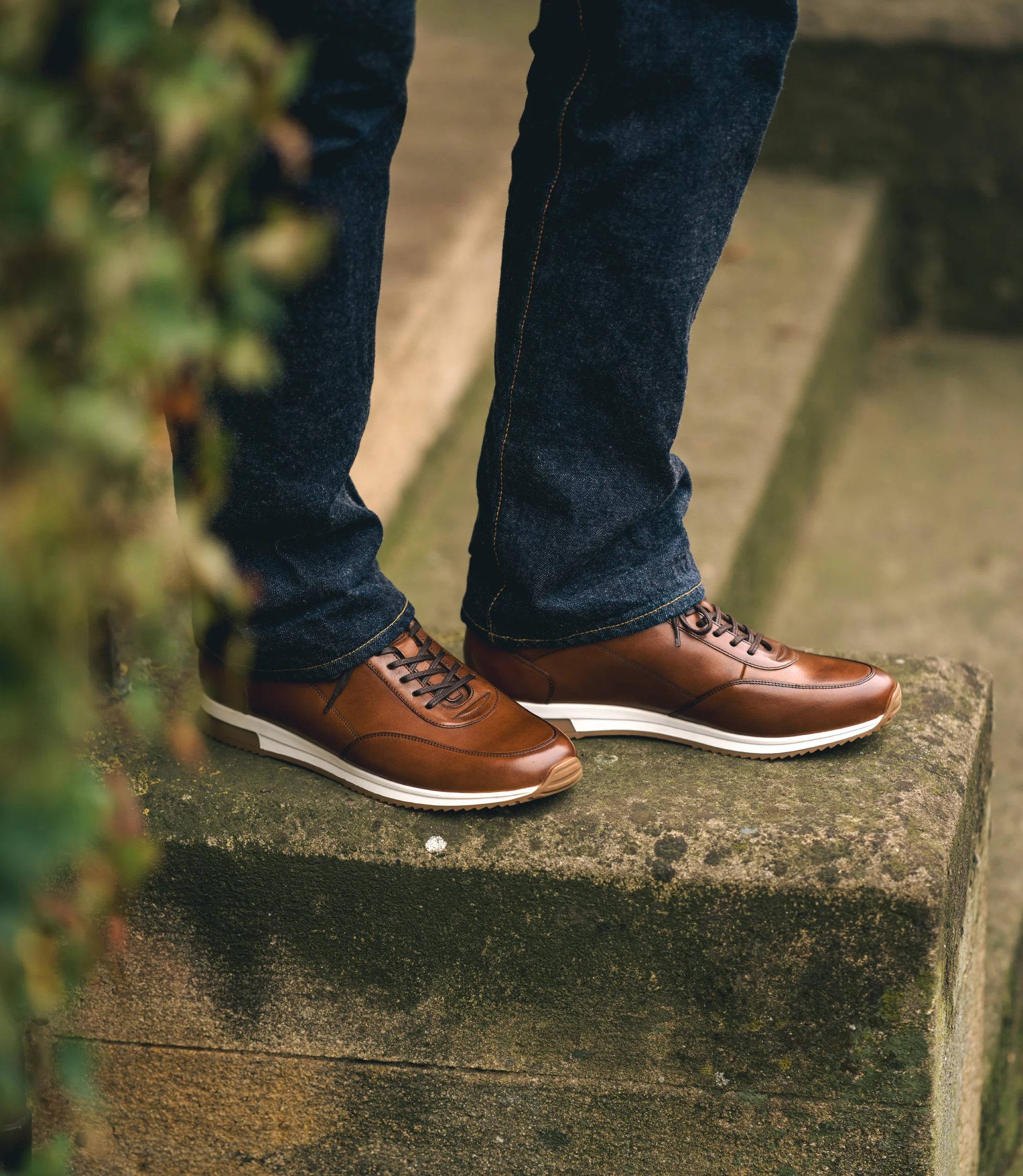 A man wearing navy denim trousers standing on a brick wall wearing his Loake Bannister Trainers in Cedar Leather.