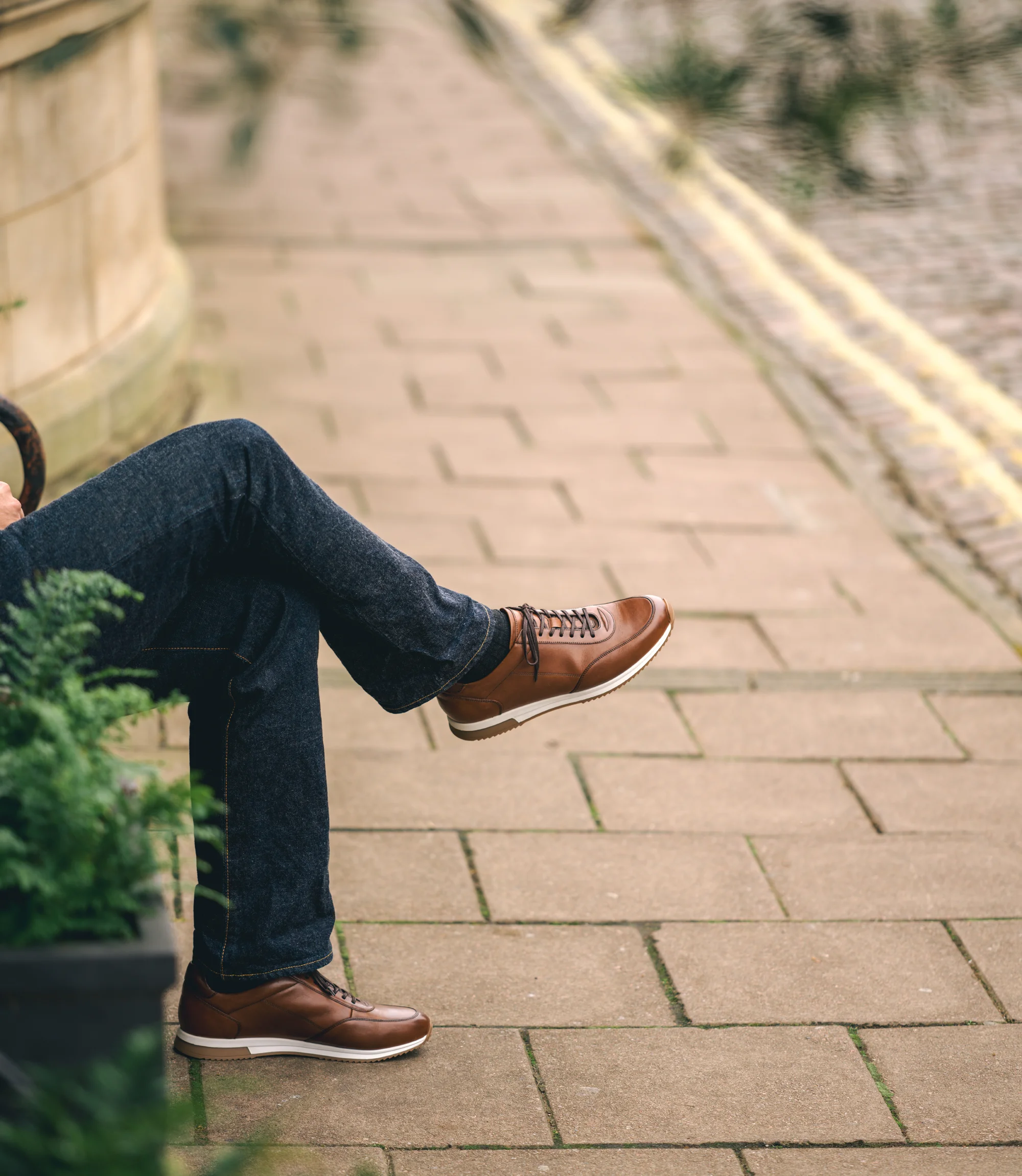 A man wearing navy denim trousers sitting on a bench legs crossed wearing his Loake Bannister Trainers in Cedar Leather.