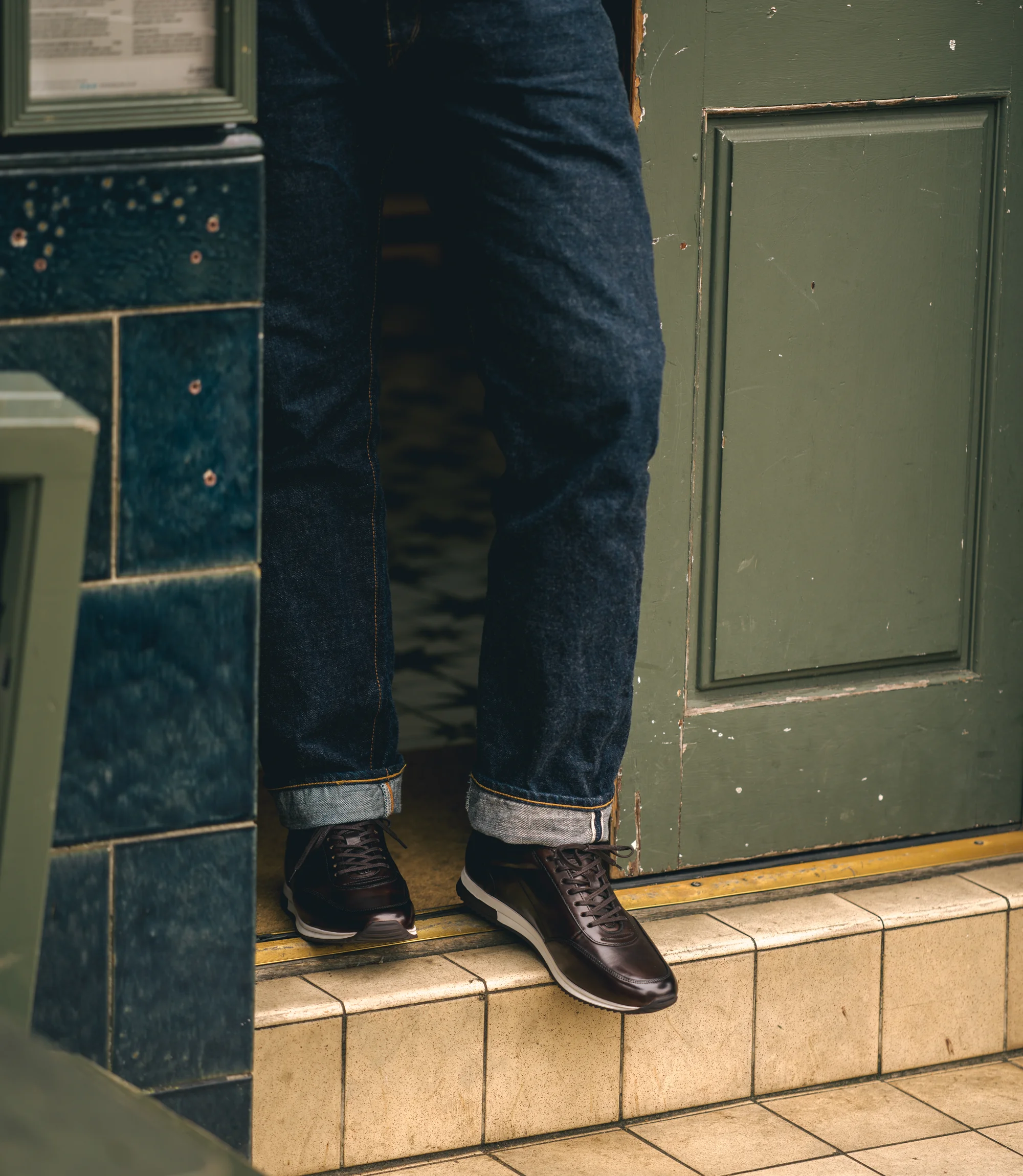 A man wearing navy denim trousers standing on a concrete step wearing his Loake Bannister Trainers in Dark Brown Leather.