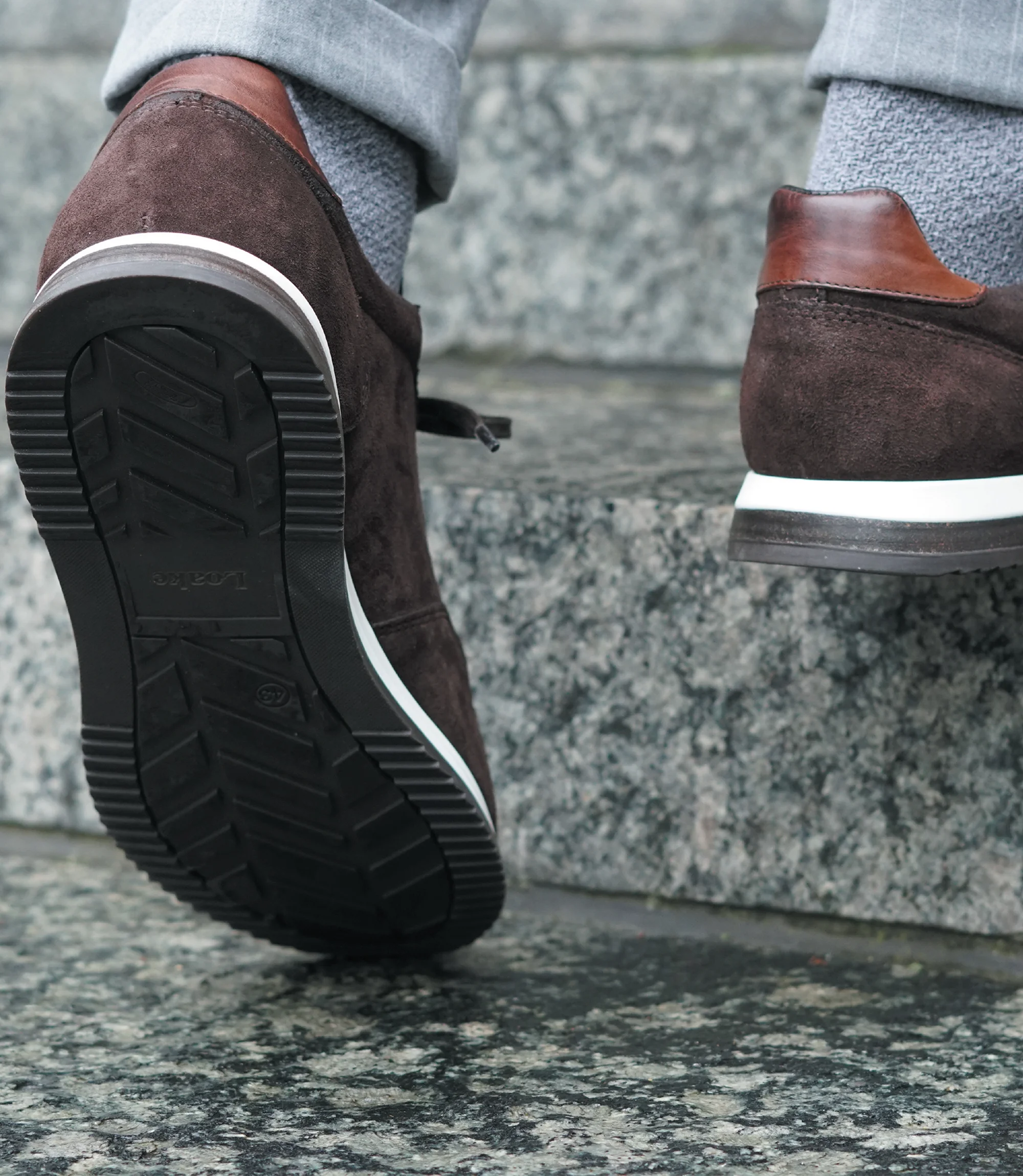 A man wearing grey trousers walking up stairs, wearing Loake Bannister Trainers in Dark Brown Suede.