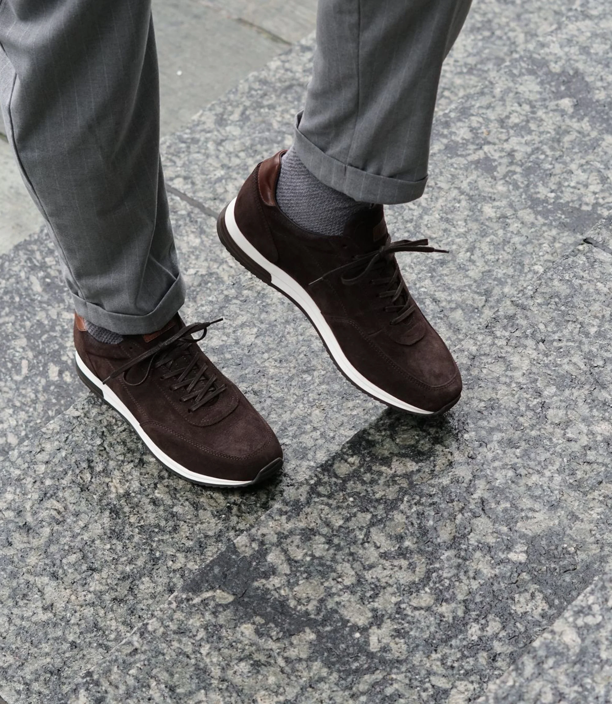 A man wearing grey trousers standing on marble floor wearing his Loake Bannister Trainers in Dark Brown Suede.