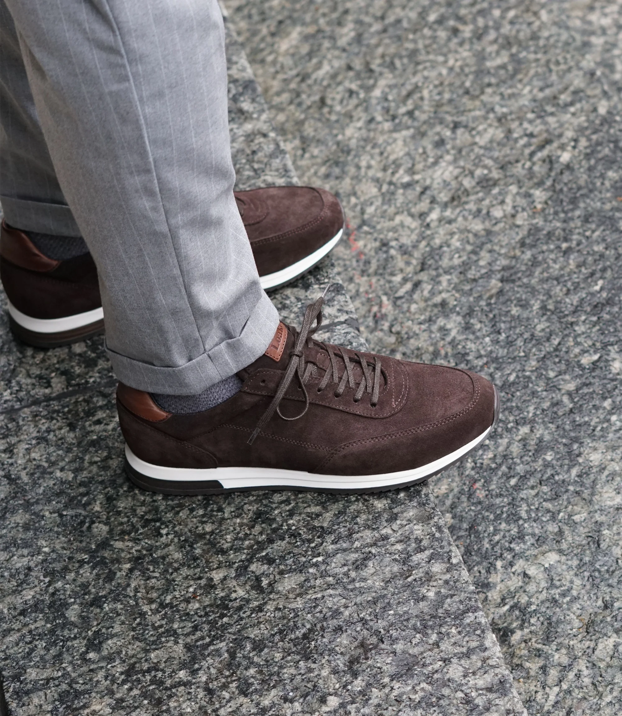 A man wearing grey trousers standing on marble floor wearing his Loake Bannister Trainers in Dark Brown Suede.