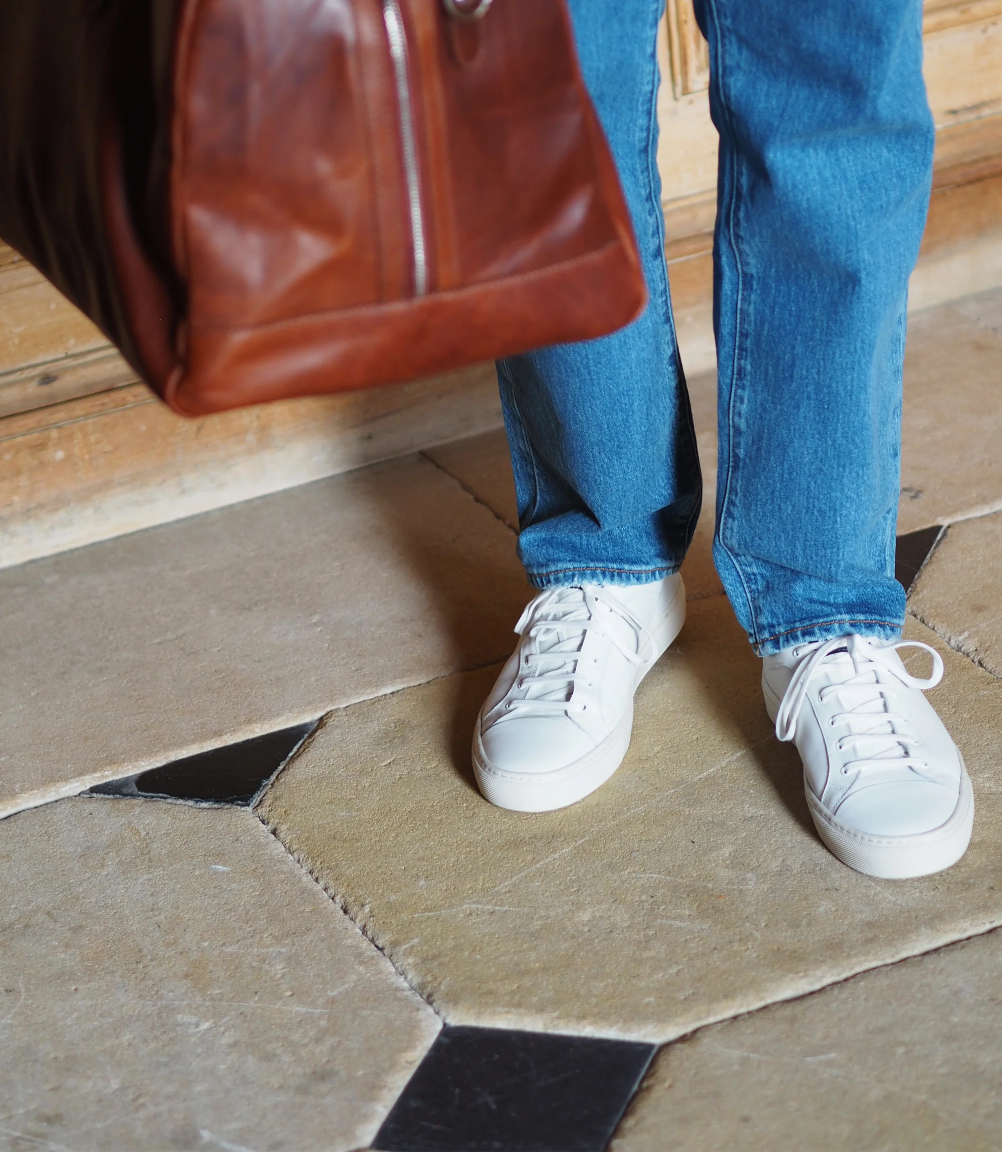 A man wearing blue denim trousers standing on concrete floor with a brown bag wearing his Loake Dash Trainers in White Leather.