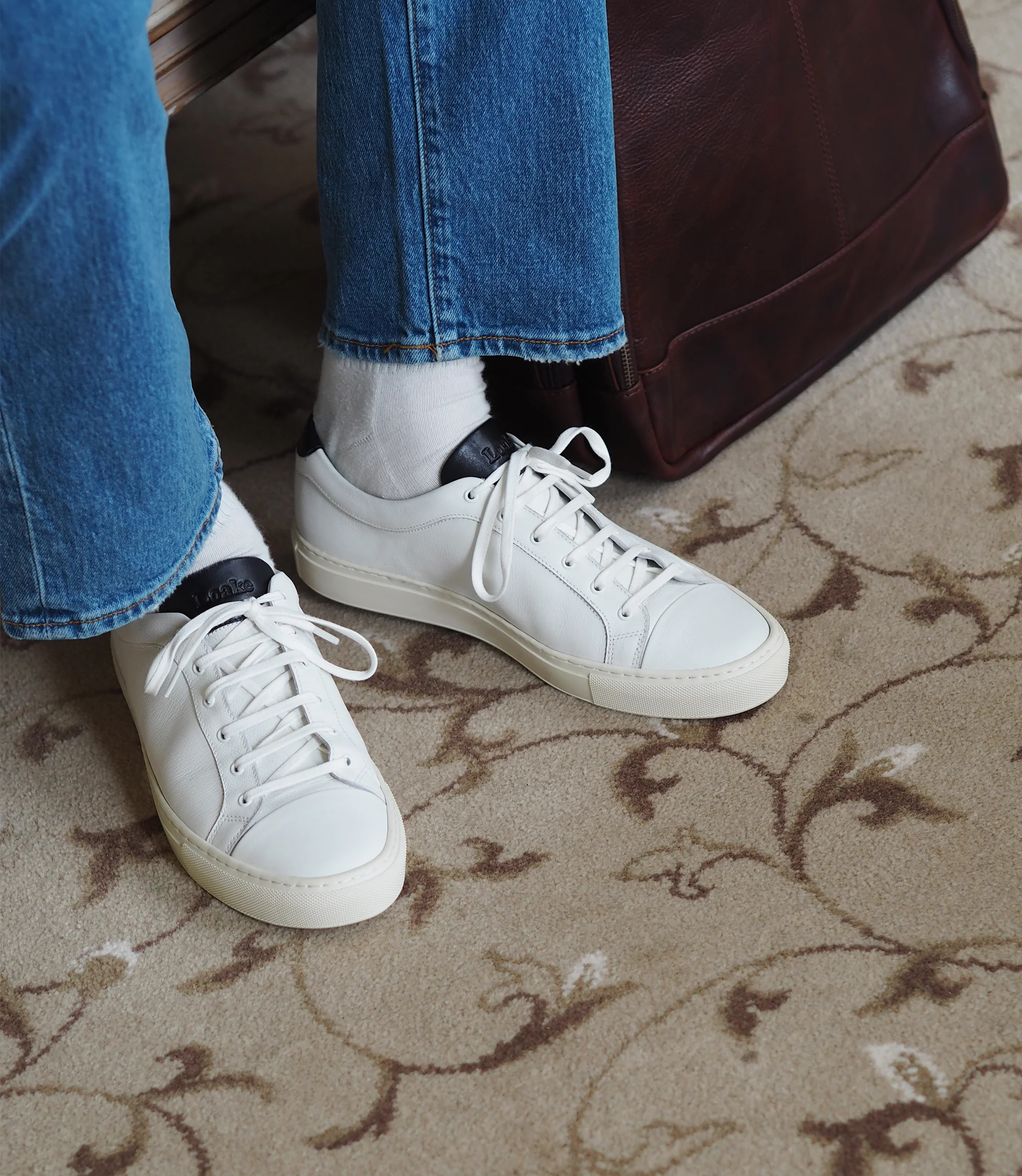 A man wearing blue denim trousers sitting on a chair in a living room with a brown rucksack wearing his Loake Dash Trainers in White Leather.