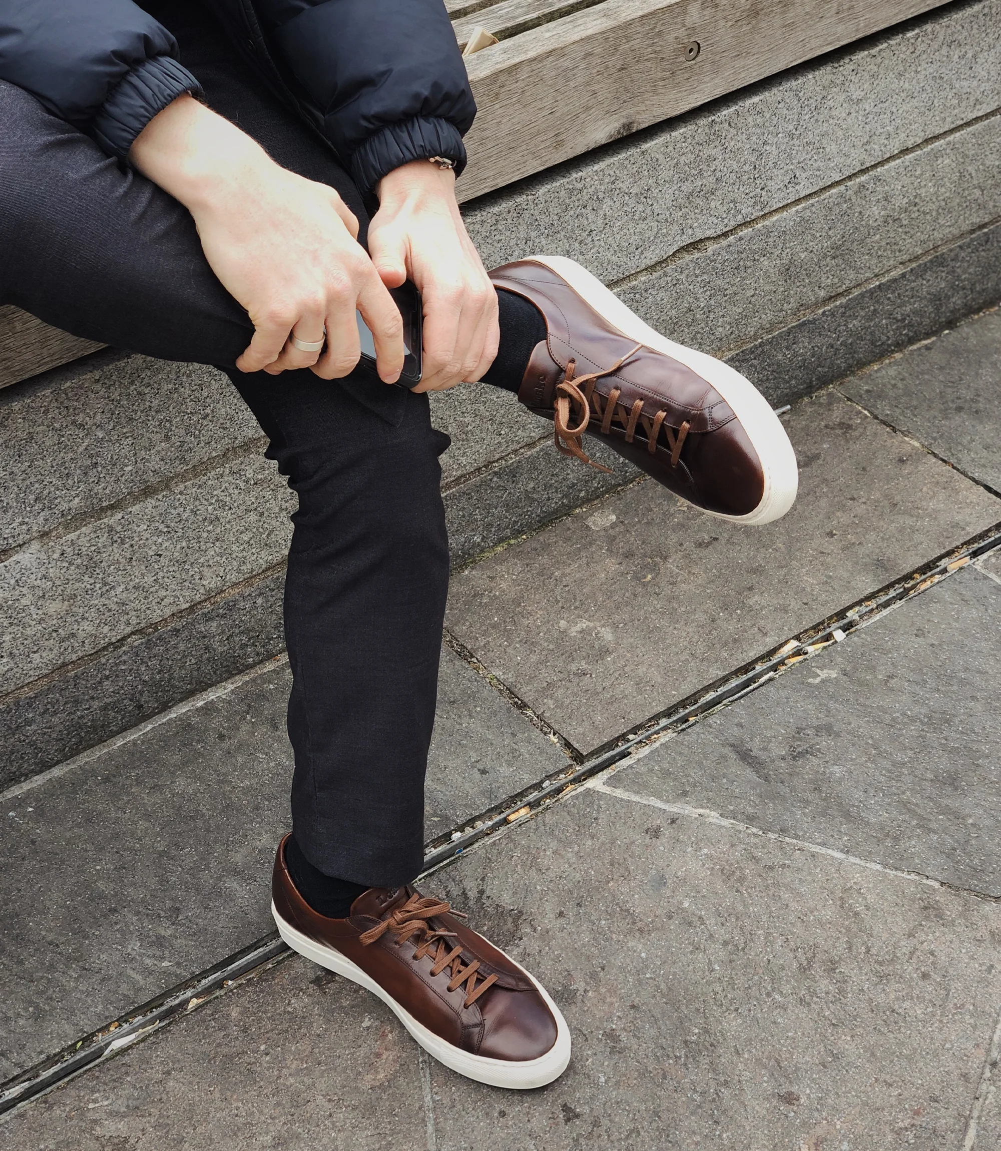 A man wearing black trousers and a navy coat sitting on a wall wearing his Loake Sprint Trainers in Dark Brown Leather.