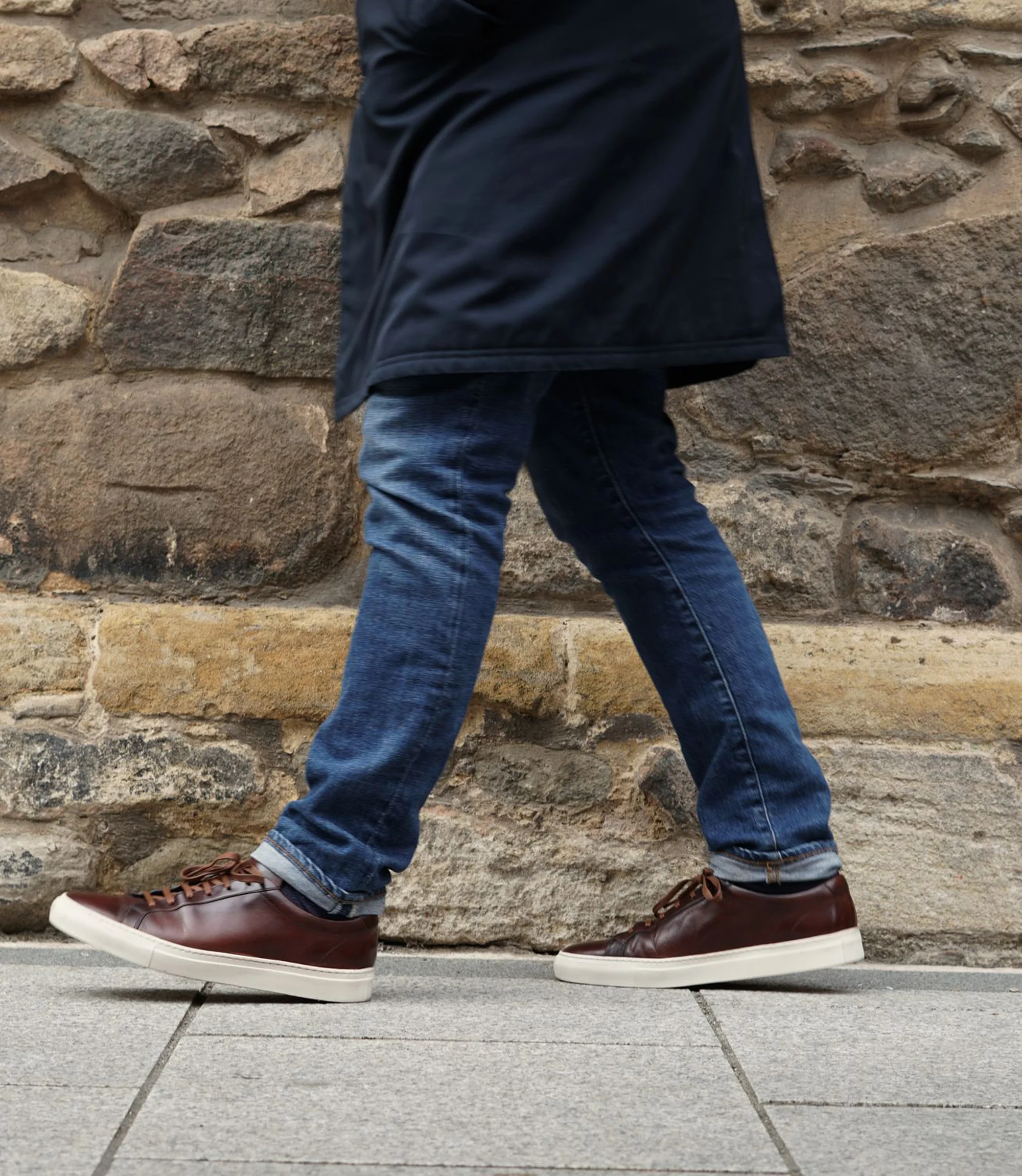 A man wearing blue denim jeans and a blue jacket walking outside in his Loake Sprint Trainers in Dark Brown Leather.