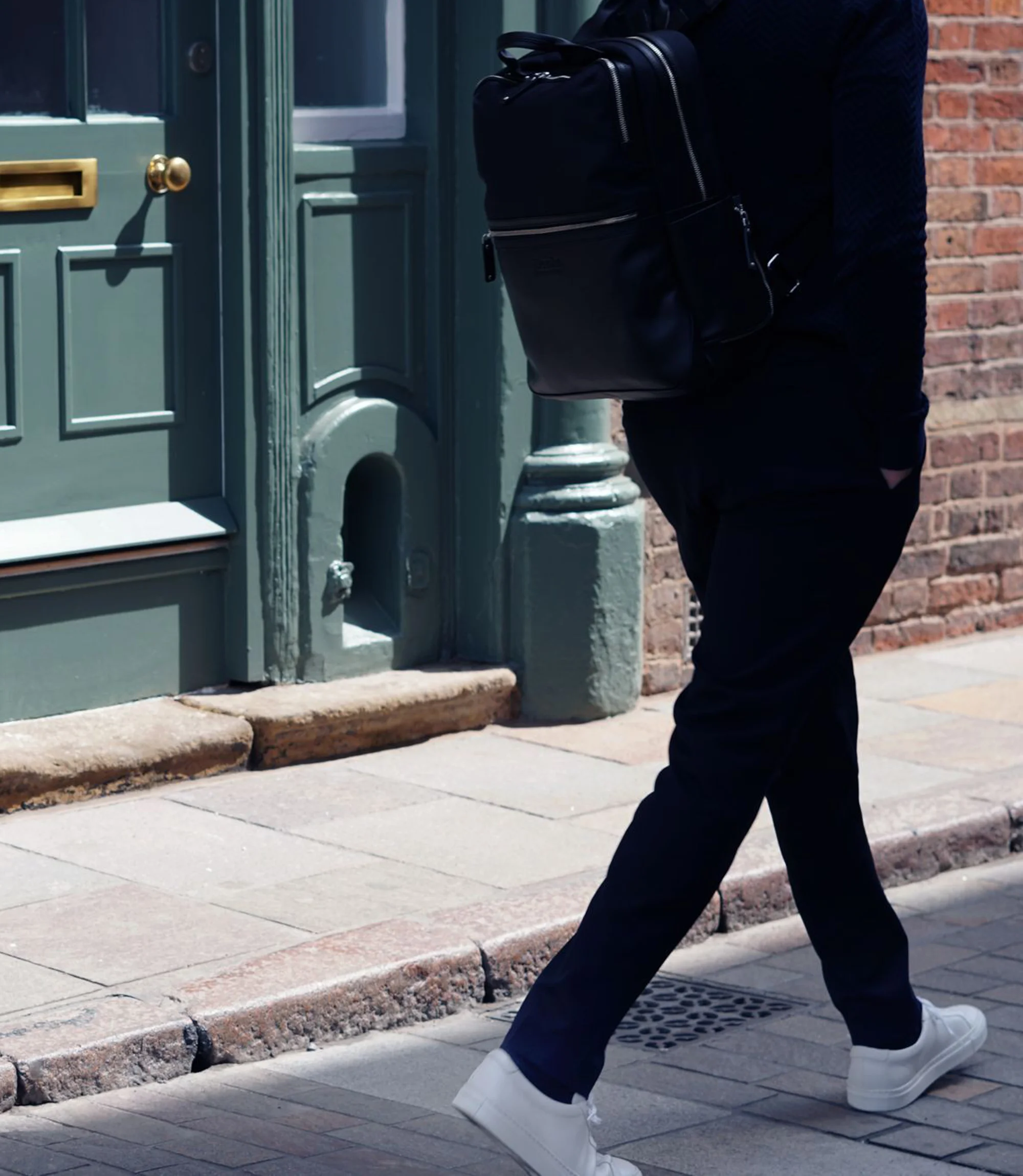 A man wearing navy trousers and a Loake Black Rucksack walking outside in his Loake Sprint Trainers in White Leather.