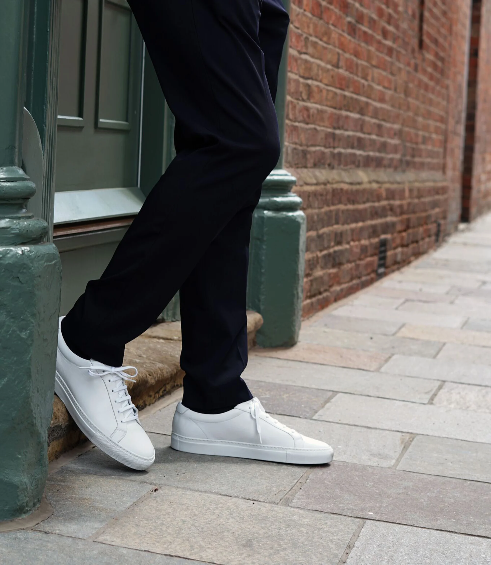 A man wearing navy trousers standing up against a green door wearing his Loake Sprint Trainers in White Leather.