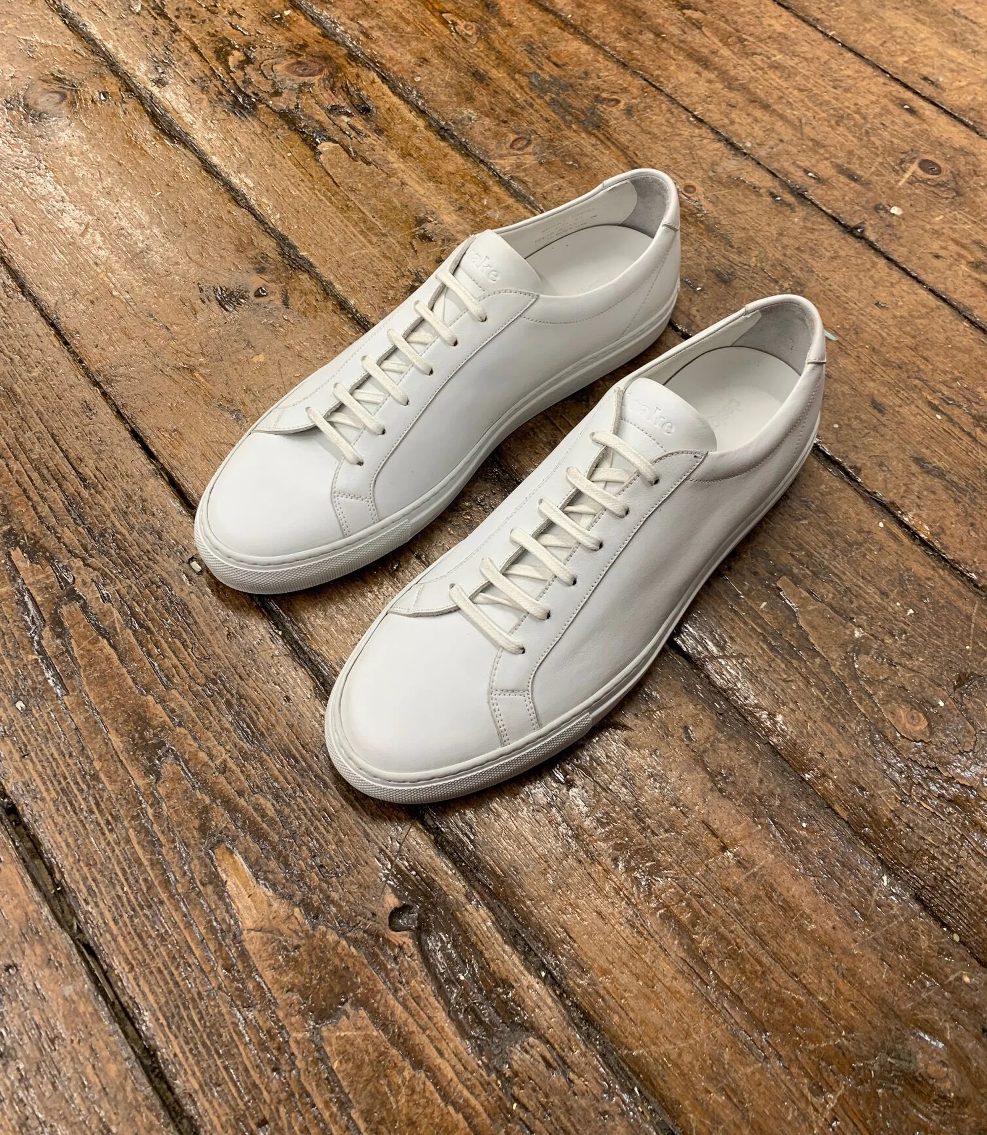 A pair of Loake Sprint Trainers in White placed on a brown wooden floor separately alone.