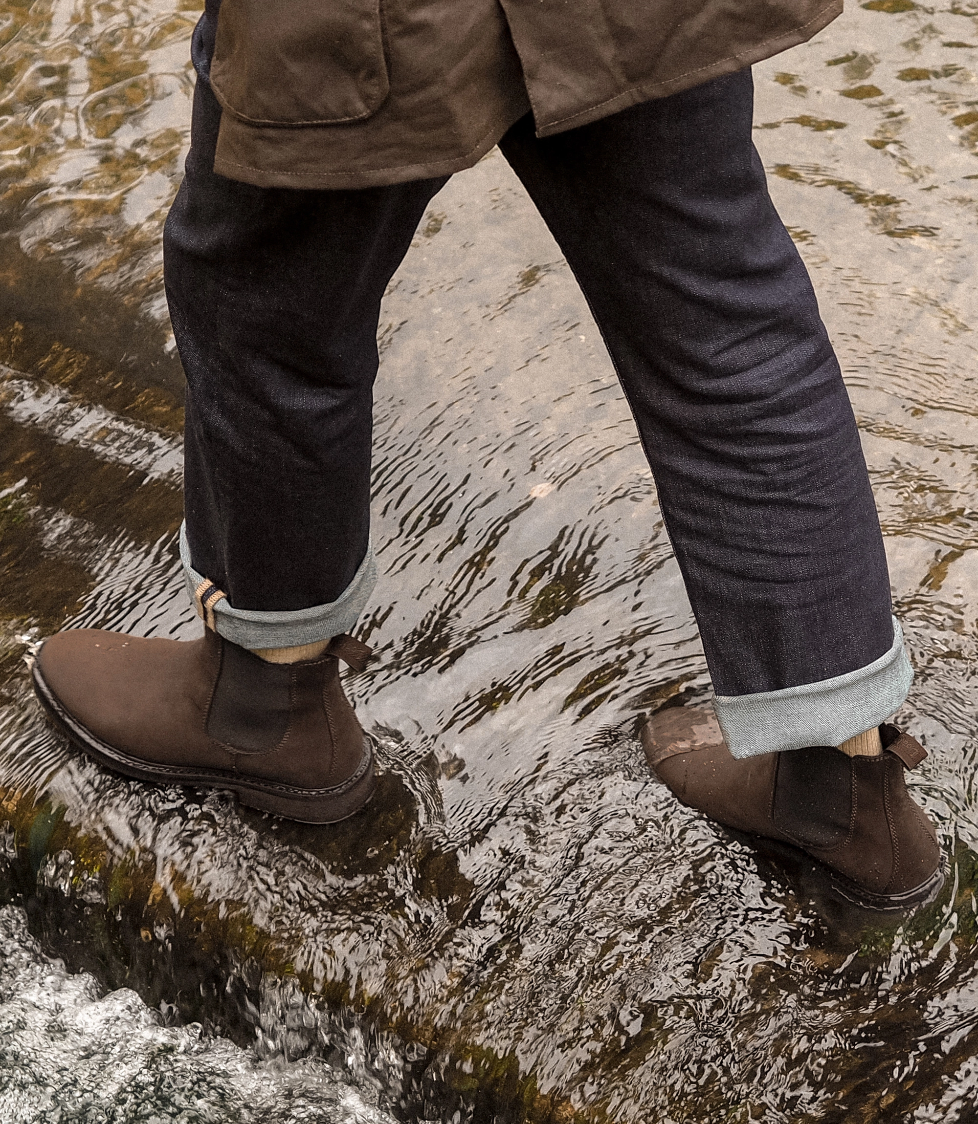 A man wearing navy jeans, brown waxy country jacket and his waterproof Ducksworth Chelsea boots. He is walking through a water ford in an English country village. Shoes shown are Ducksworth in Brown Oiled Nubuck leather