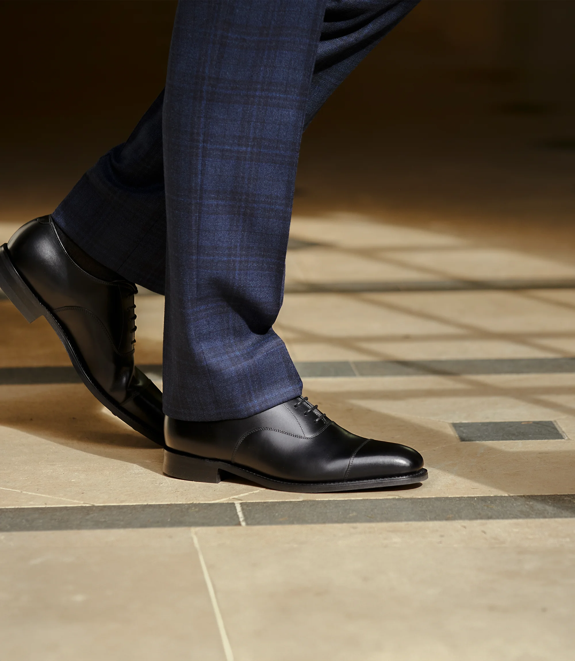 A man wearing navy suit trousers, walking on a tiled floor in their Goodyear welted leather soled Toecap Derby shoes. Shoes shown are Aldwych in Black calf leather
