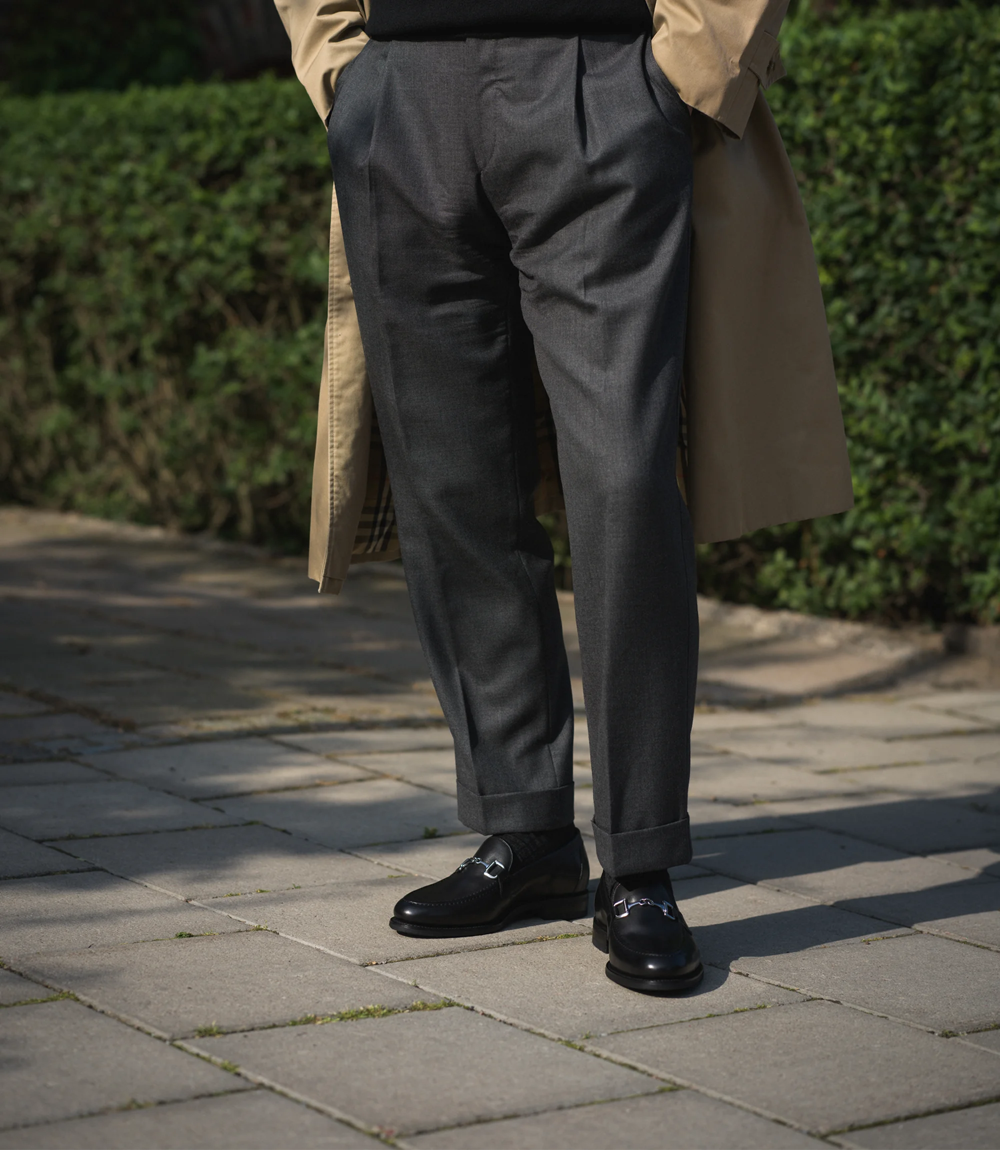 A man in a beige coat wearing his grey trousers standing on the pavement wearing his Loake Loafers Kensington in Black Leather.