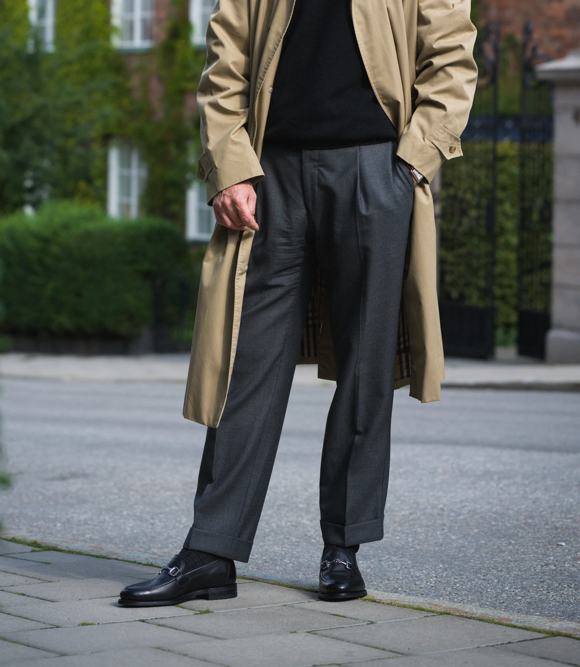 A man in a beige coat wearing his grey trousers standing on the pavement wearing his Loake Loafers Kensington in Black Leather.
