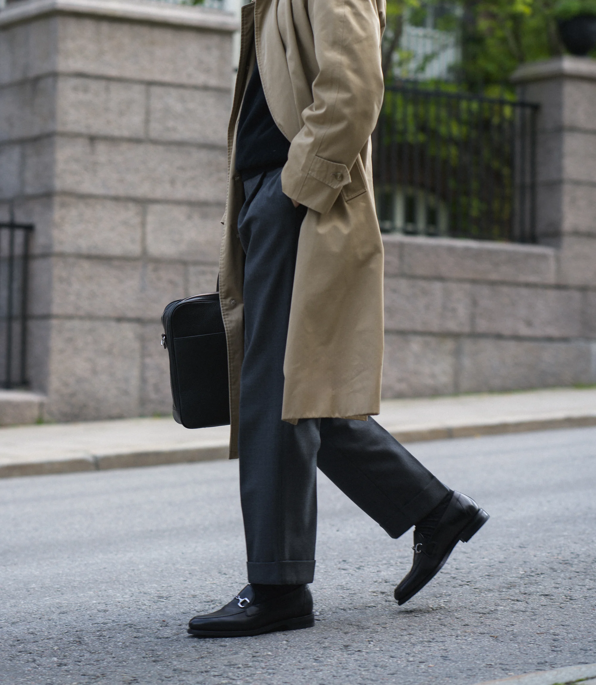 A man in a beige coat wearing his grey trousers walking across the road holding his Loake black bag wearing his Loake Loafers Kensington in Black Leather.