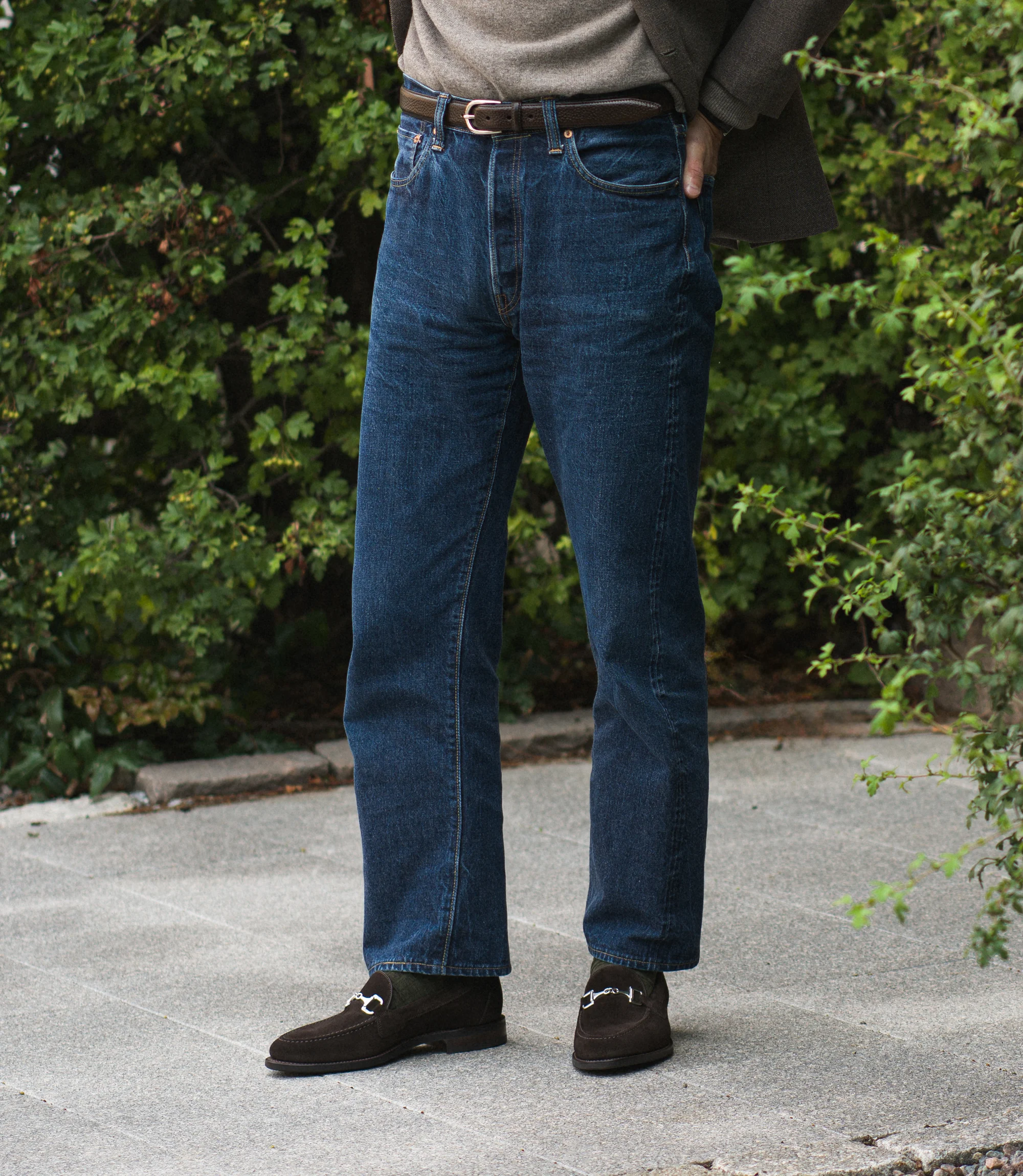 A man wearing navy denim jeans standing on concrete floor outside wearing his Loake Loafers Kensington in Dark Brown Suede.