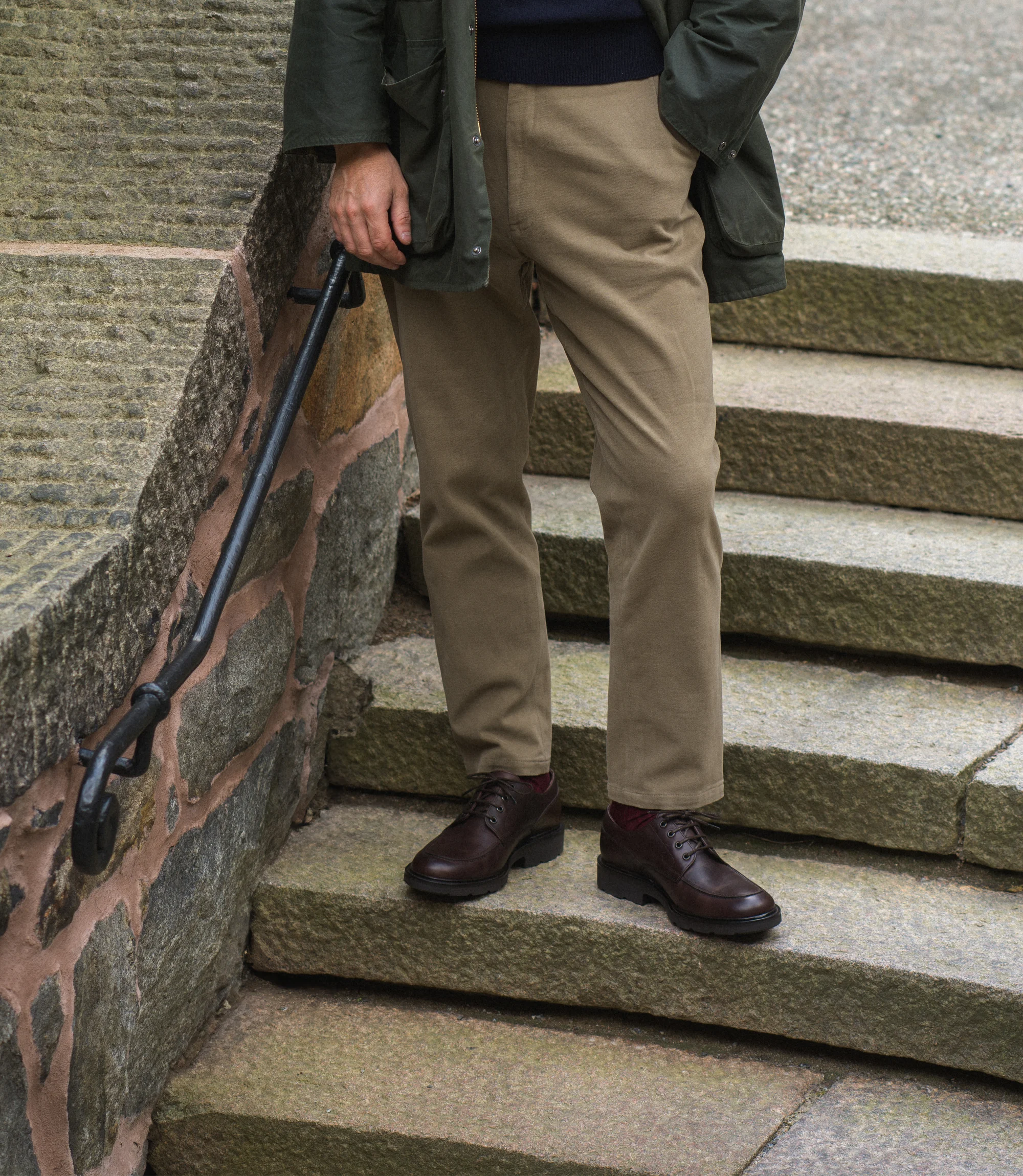 A man wearing beige trousers and green coat standing on concrete steps wearing his Loake Mowbury in Dark Brown leather.