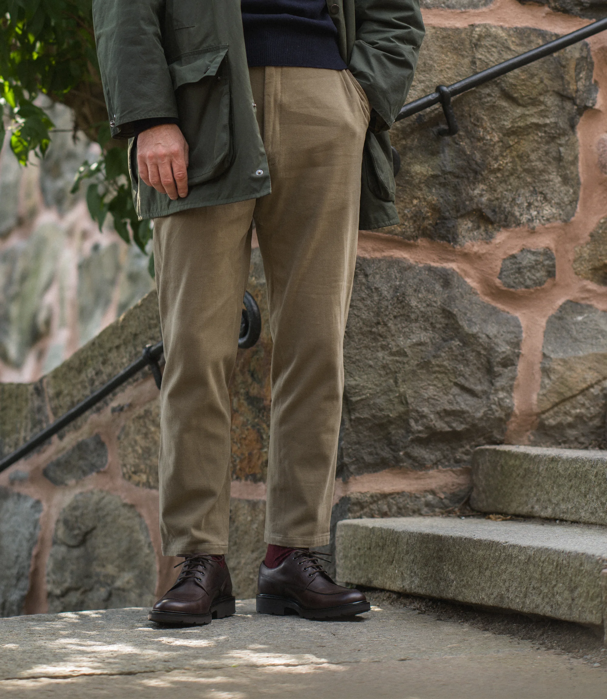 A man wearing beige trousers and green coat standing outside near steps wearing his Loake Mowbury in Dark Brown leather.