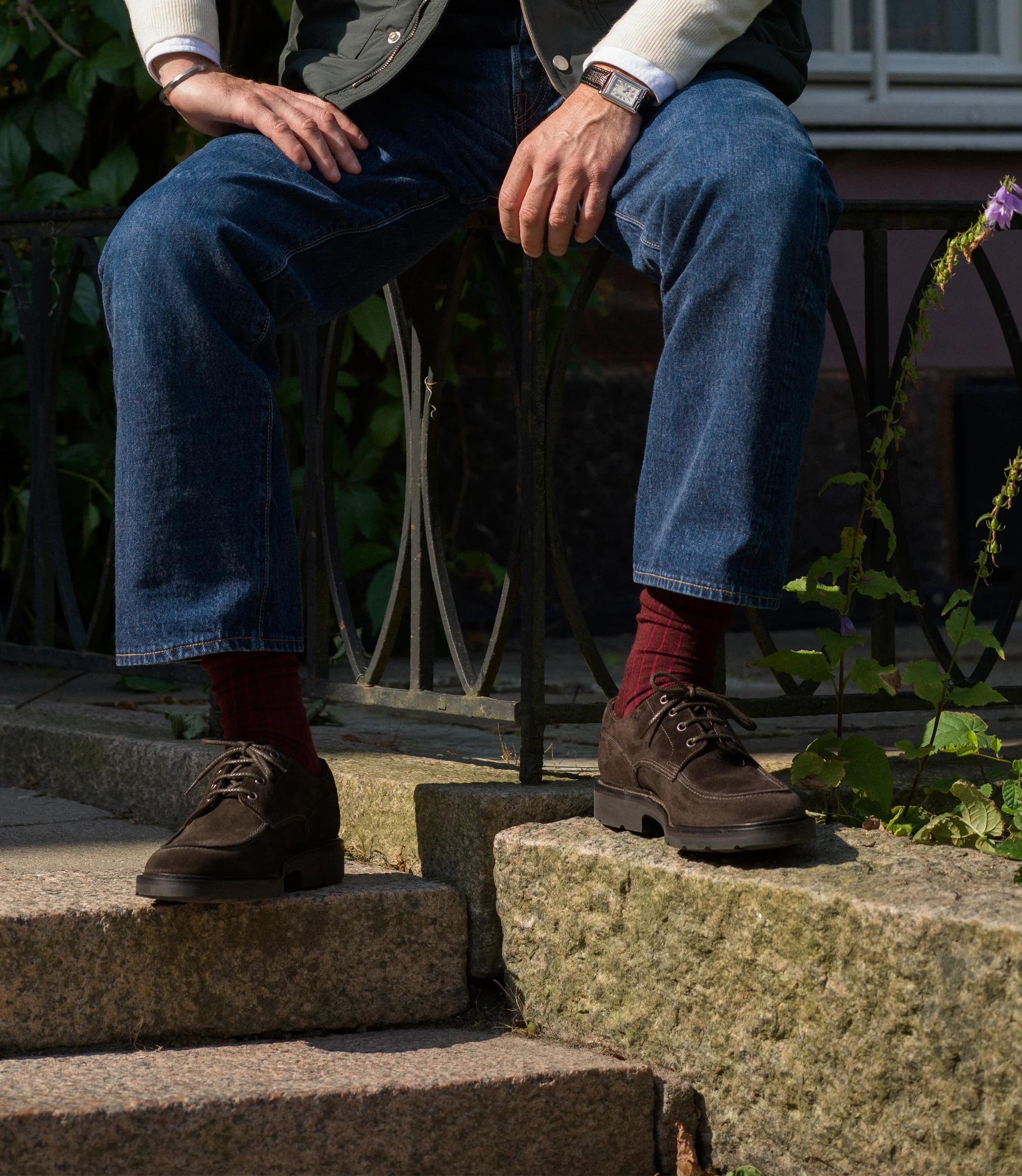 A man wearing navy denim jeans sitting on a fence outside wearing his Loake Mowbury in Dark Brown Suede.