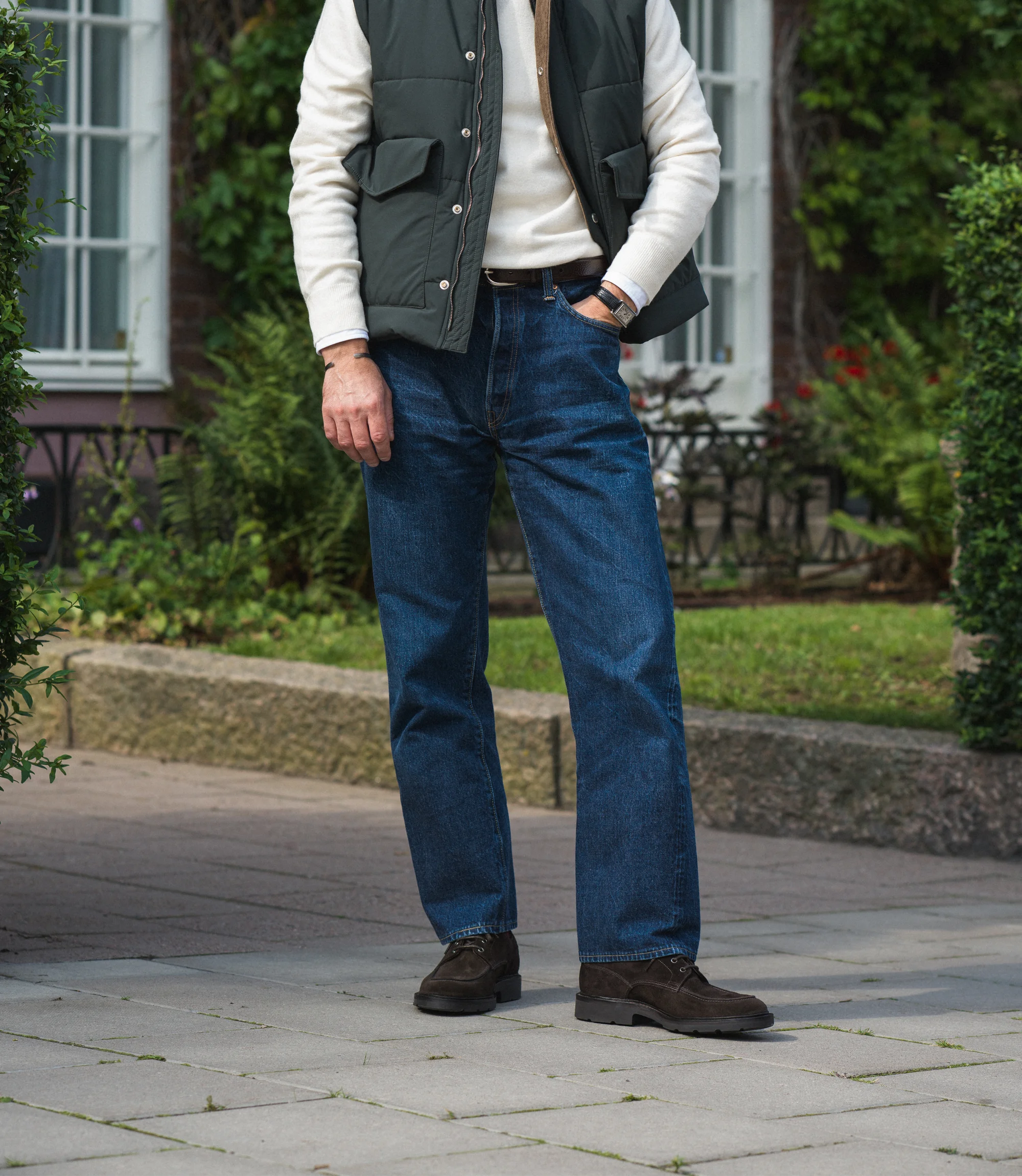 A man wearing navy denim jeans and a green gilet standing outside wearing his Loake Mowbury in Dark Brown Suede.
