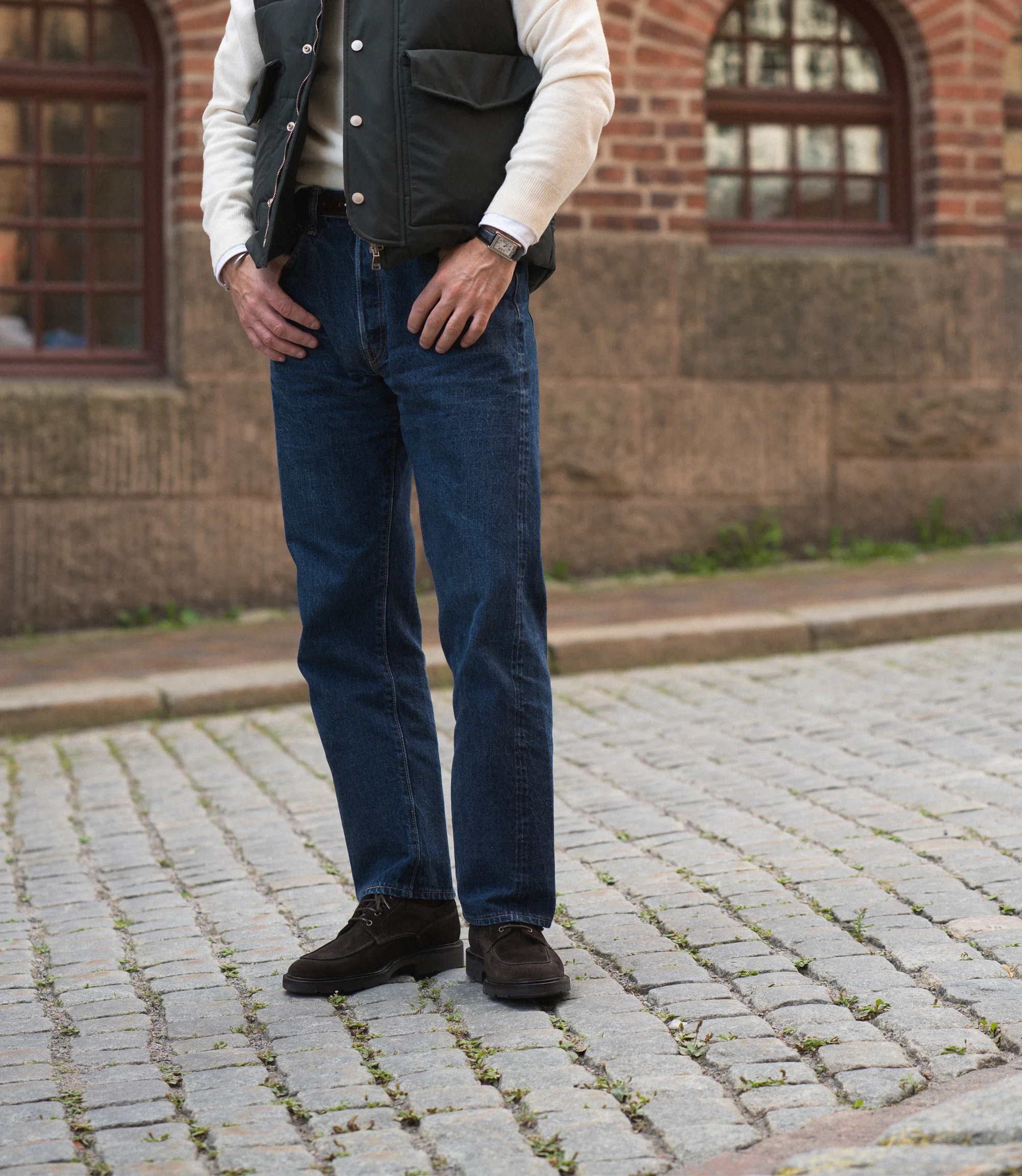A man wearing navy denim jeans and a green gilet standing outside on concrete flooring wearing his Loake Mowbury in Dark Brown Suede.