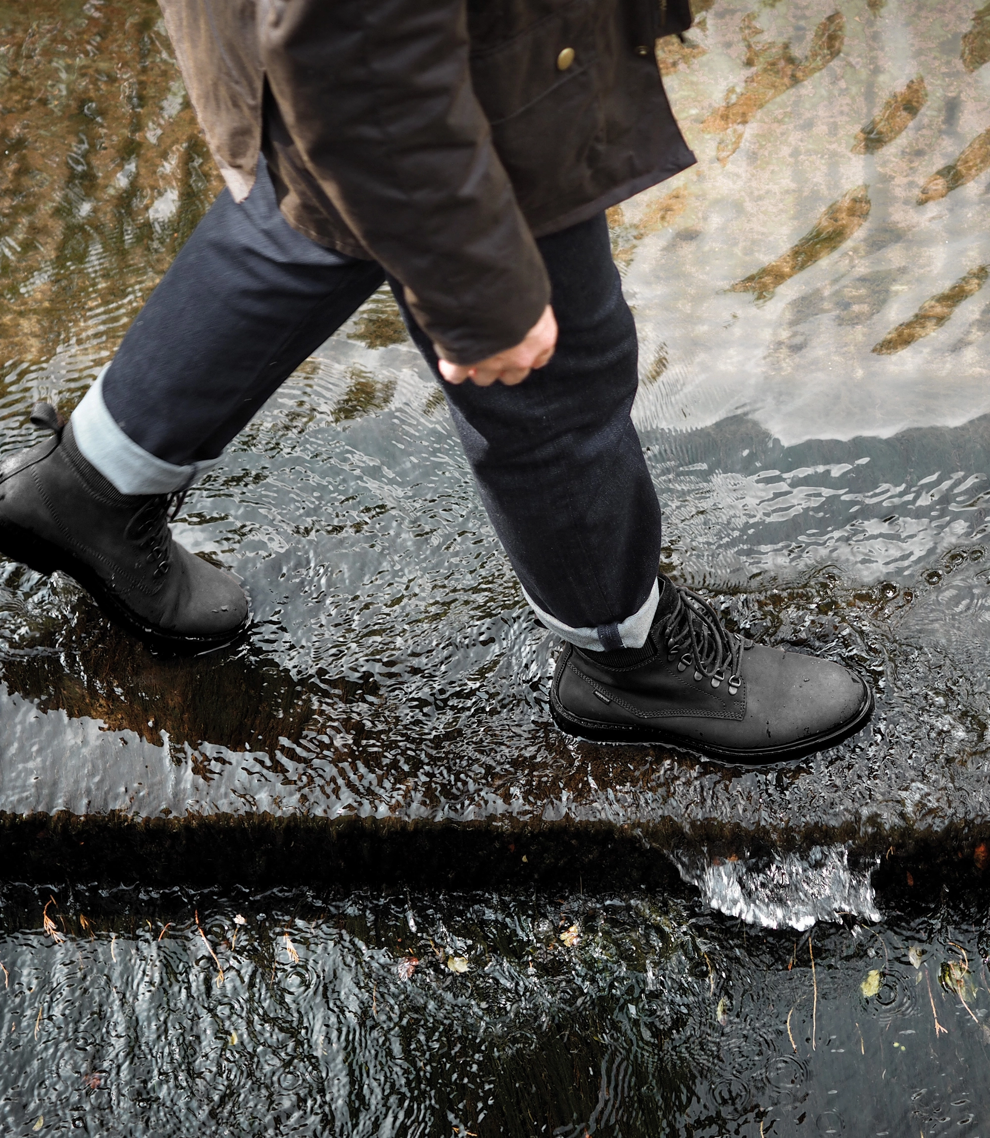 A man wearing navy blue denim trousers, brown waxy country jacket and his waterproof Loake Nevis Derby Boots. He is walking through a water ford in an English country village. Boots shown are Nevis in Black Oiled Nubuck leather