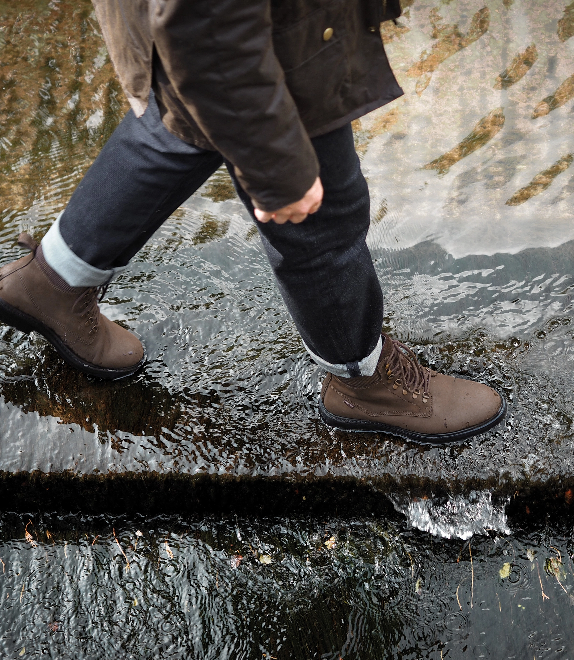 A man wearing navy blue denim trousers, brown waxy country jacket and his waterproof Loake Nevis Derby Boots. He is walking through a water ford in an English country village. Boots shown are Nevis in Brown Oiled Nubuck leather