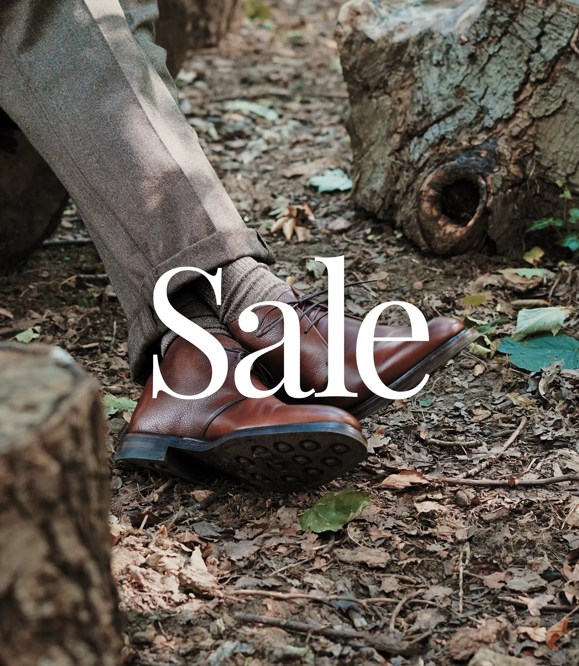 Man wearing beige trousers sitting on a tree stump in a wooded area, wearing their Loake chukka boots. Shoes shown are Lytham Burgundy Grain