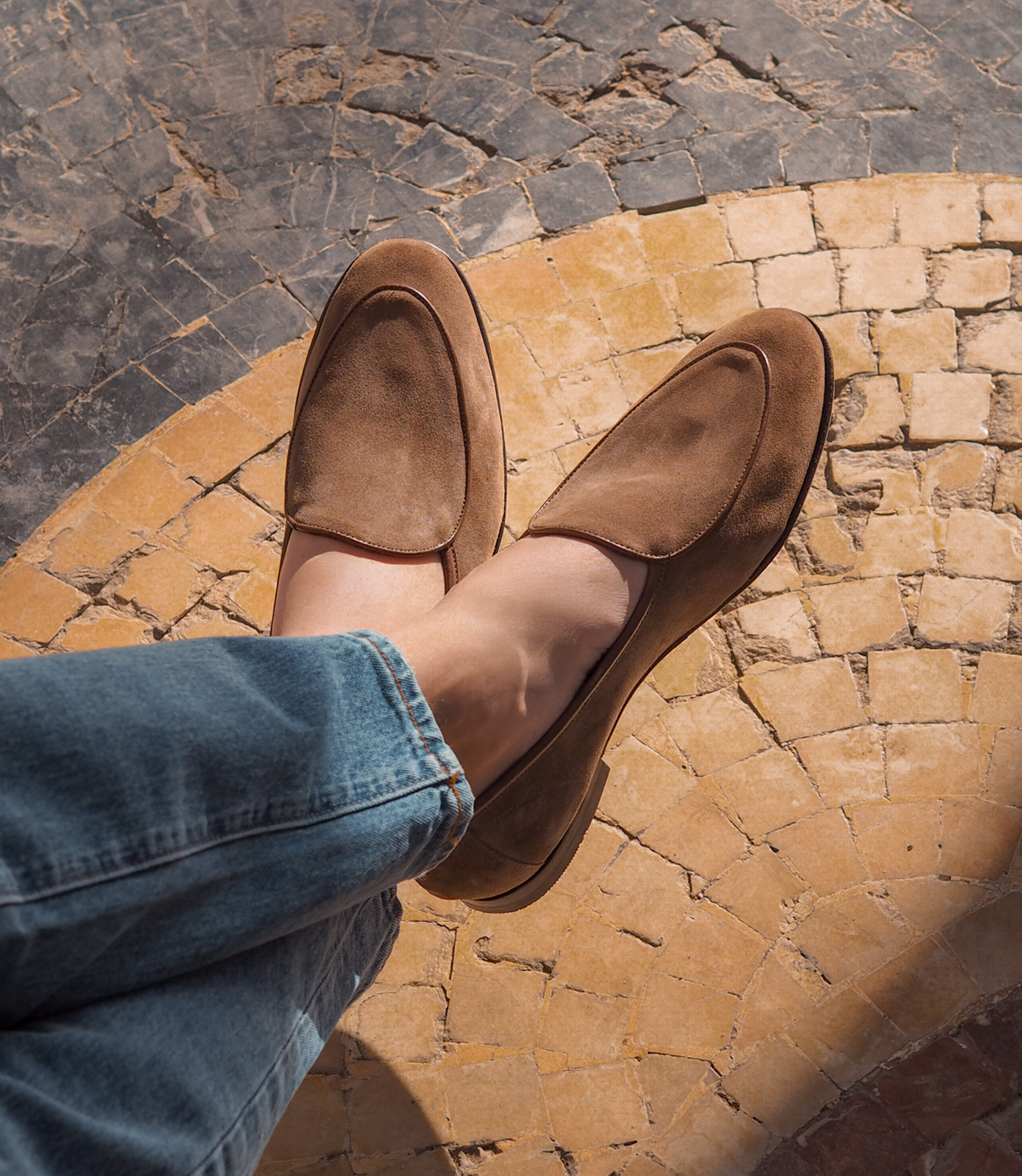 A man wearing light navy blue jeans sitting down with legs crossed on holiday wearing his Loake Belgrave Loafer in Chestnut Suede.