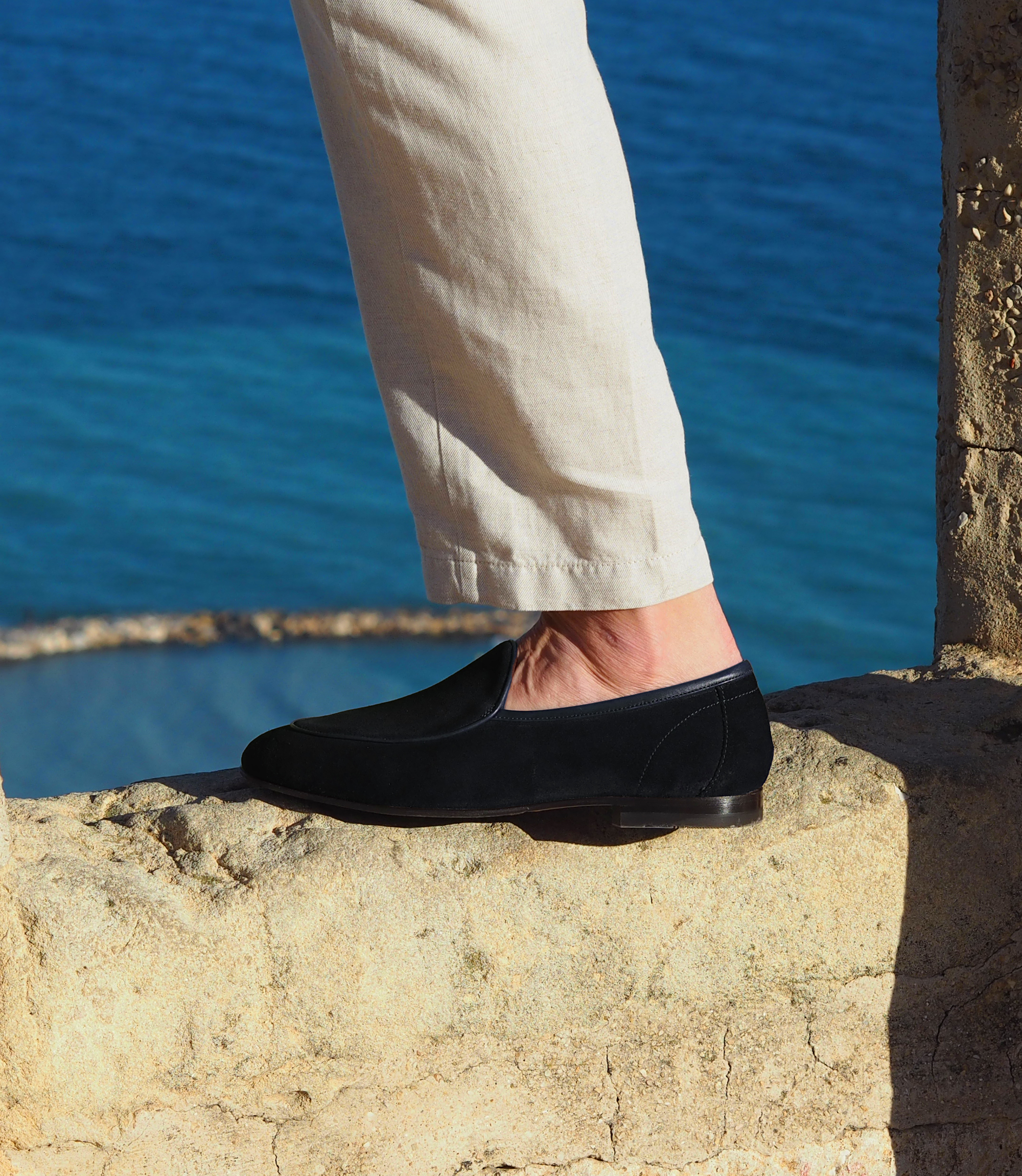A man standing outside with the sea in the background wearing a navy shirt and white linen trousers wearing his Loake Belgrave Loafers in navy suede.