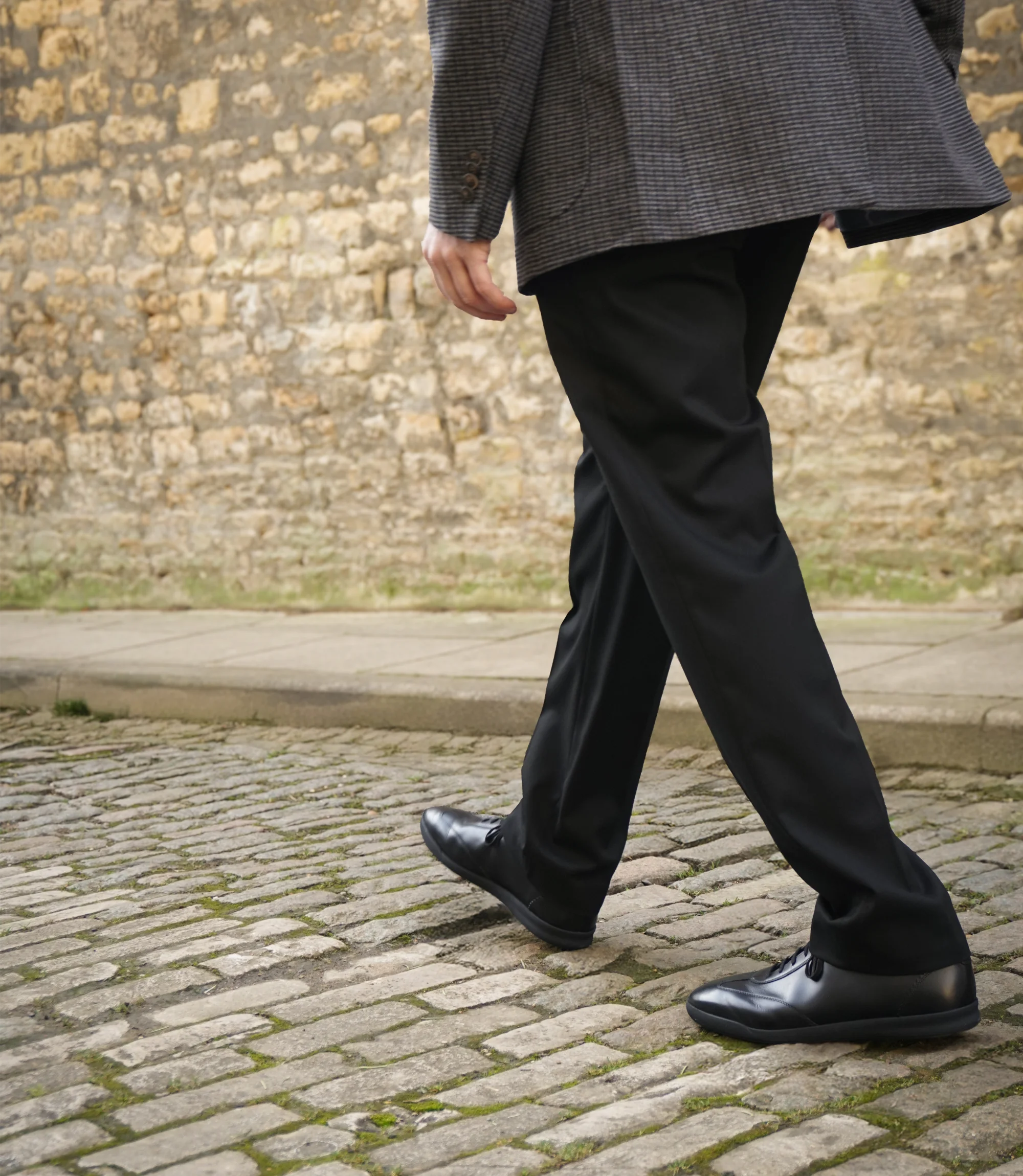 A man walking on cobbled street in black smart trousers wearing his Loake Mayfair Smart Trainers in Black.