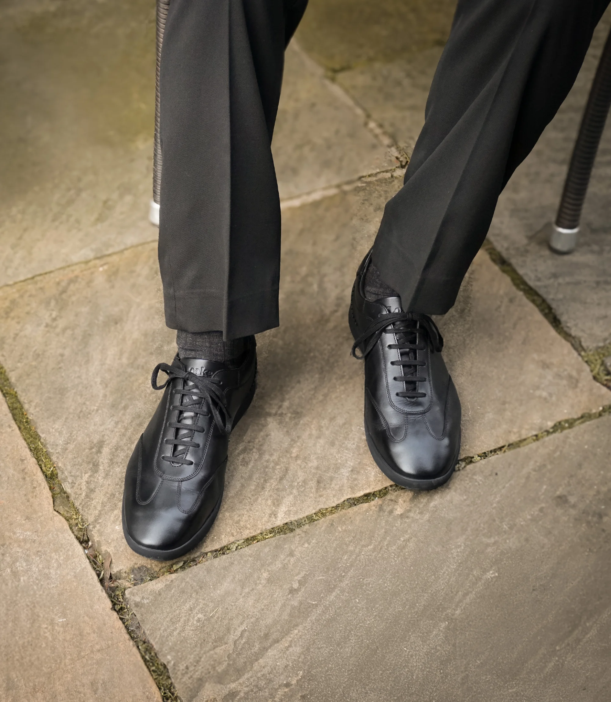 A man sitting down in the street in black smart trousers wearing his Loake Mayfair Smart Trainers in Black.
