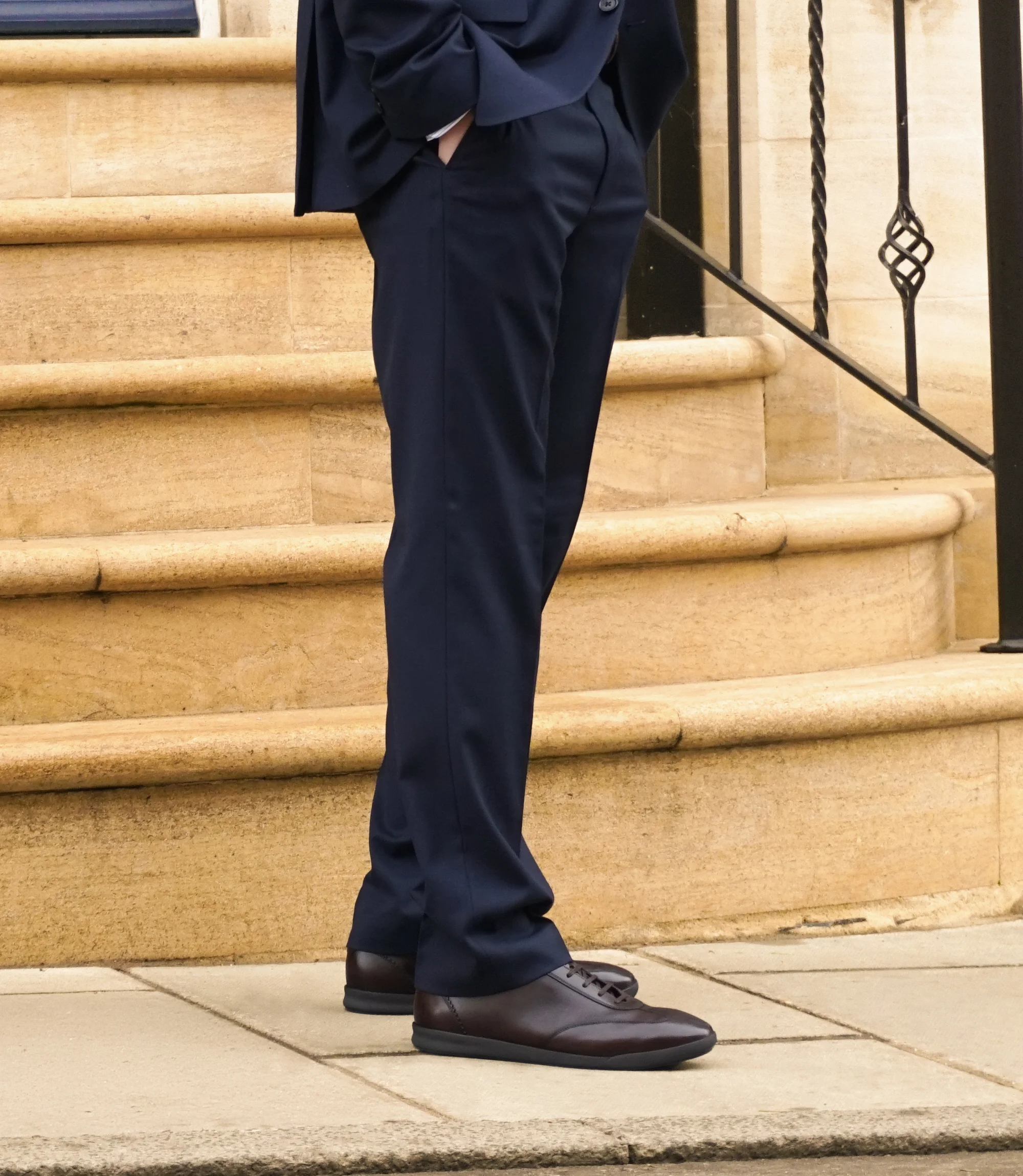A man in a navy blue suit standing in the street wearing his Loake Mayfair Smart Trainers in Dark Brown Leather.