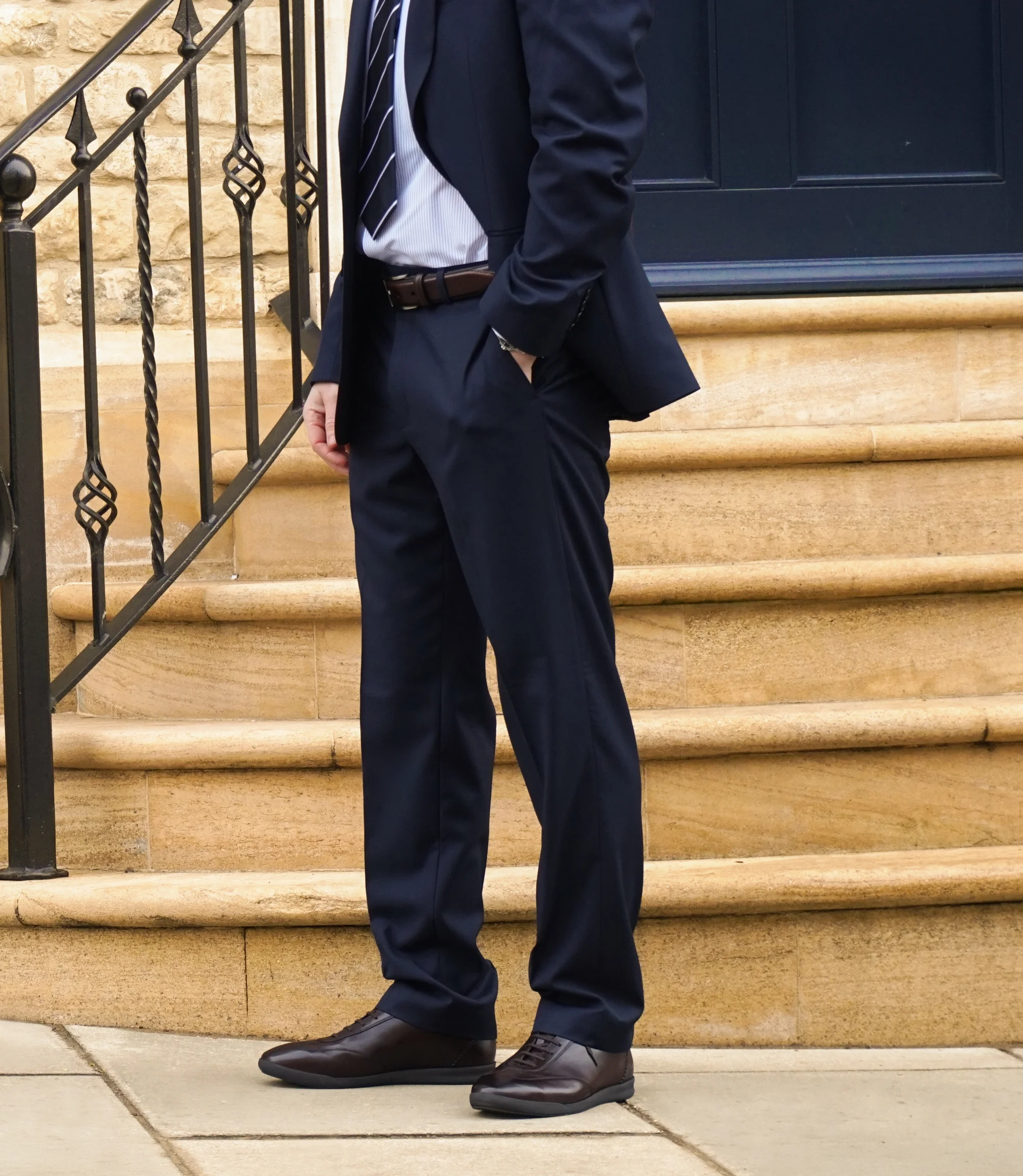 A man in a navy blue suit standing in the street wearing his Loake Mayfair Smart Trainers in Dark Brown Leather.