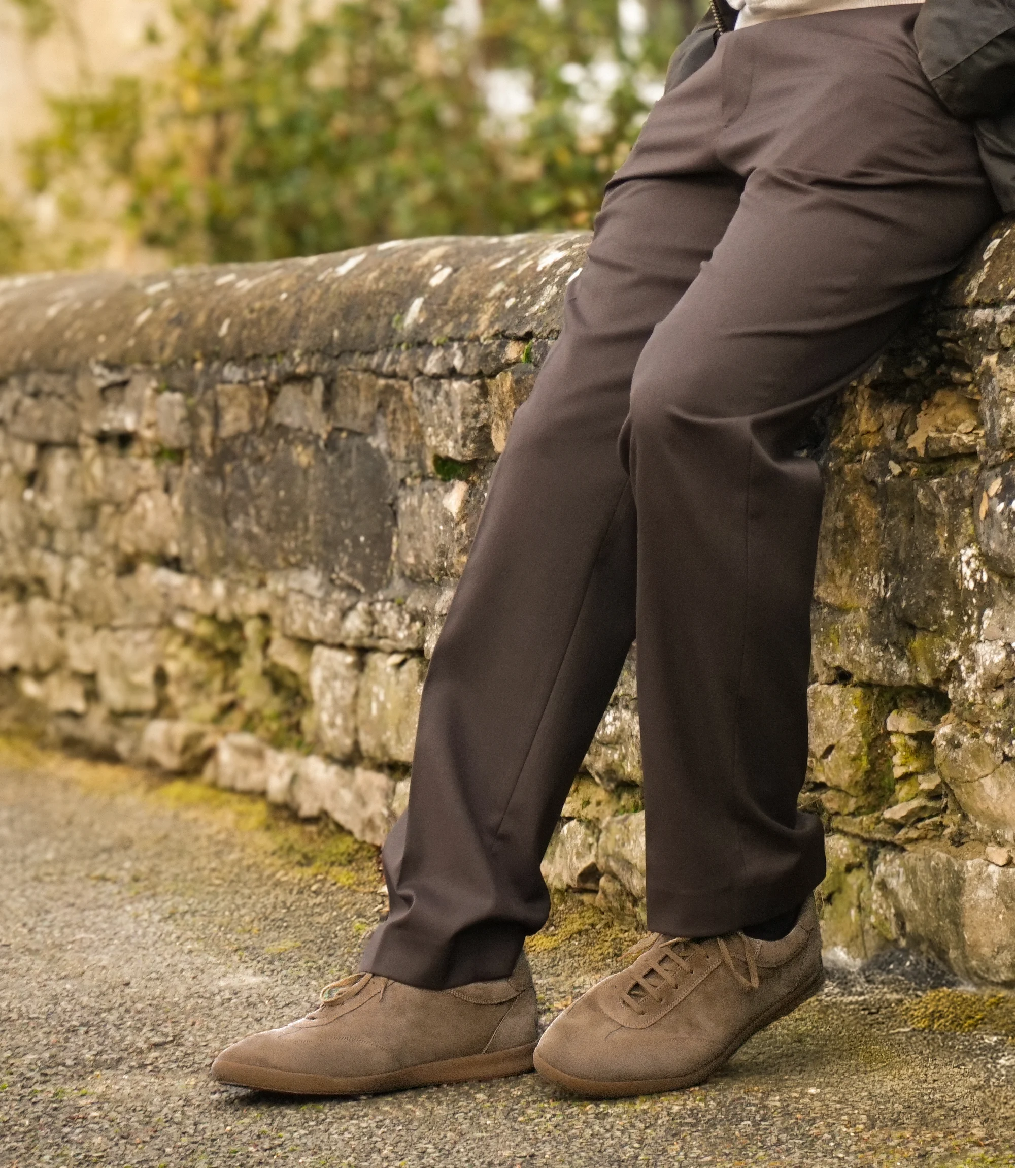 A man in smart brown trousers and brown coat standing in the street wearing his Loake Mayfair Smart Trainers in Flint Suede.