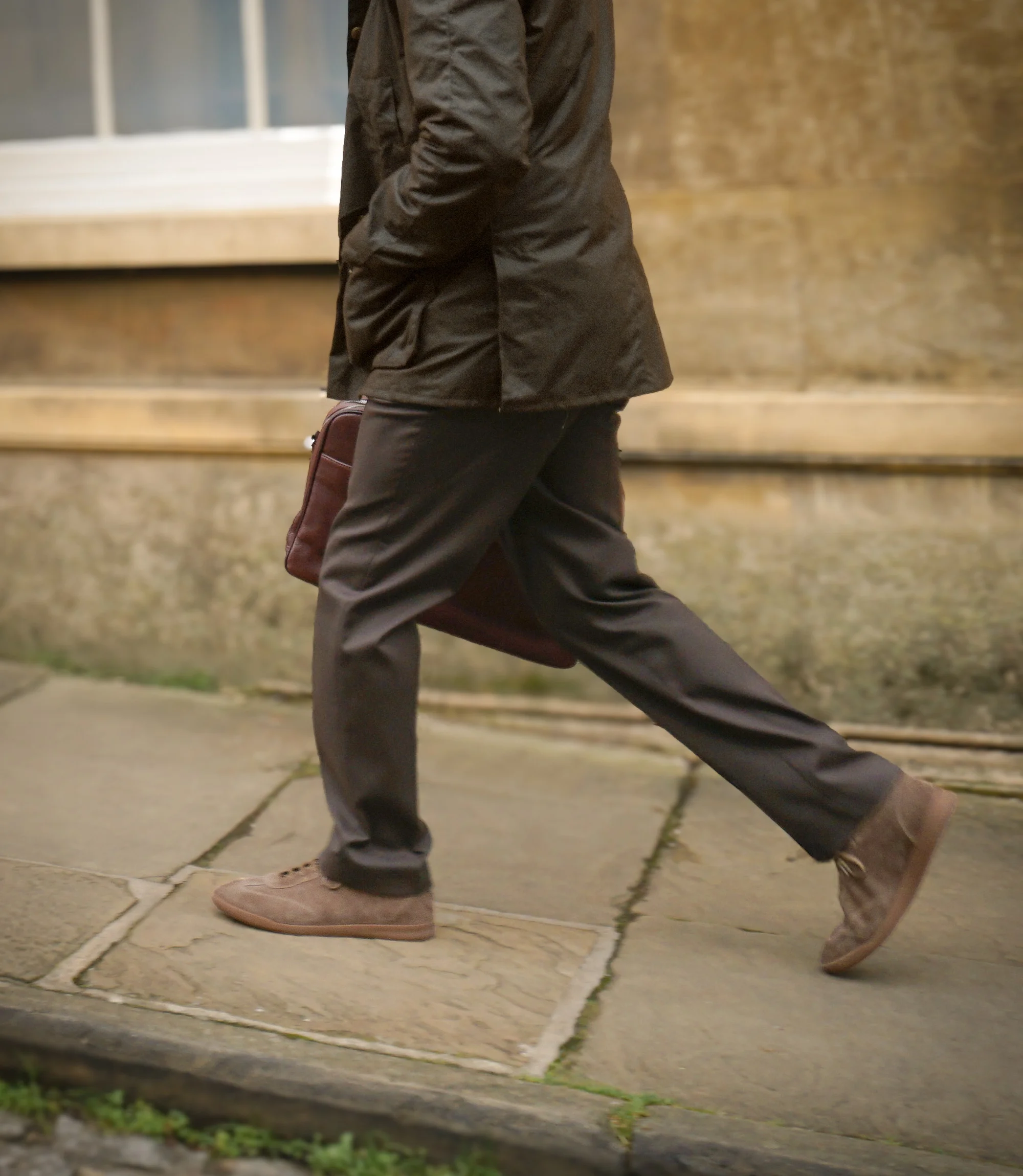 A man in smart brown trousers and brown coat walking in the street wearing his Loake Mayfair Smart Trainers in Flint Suede.