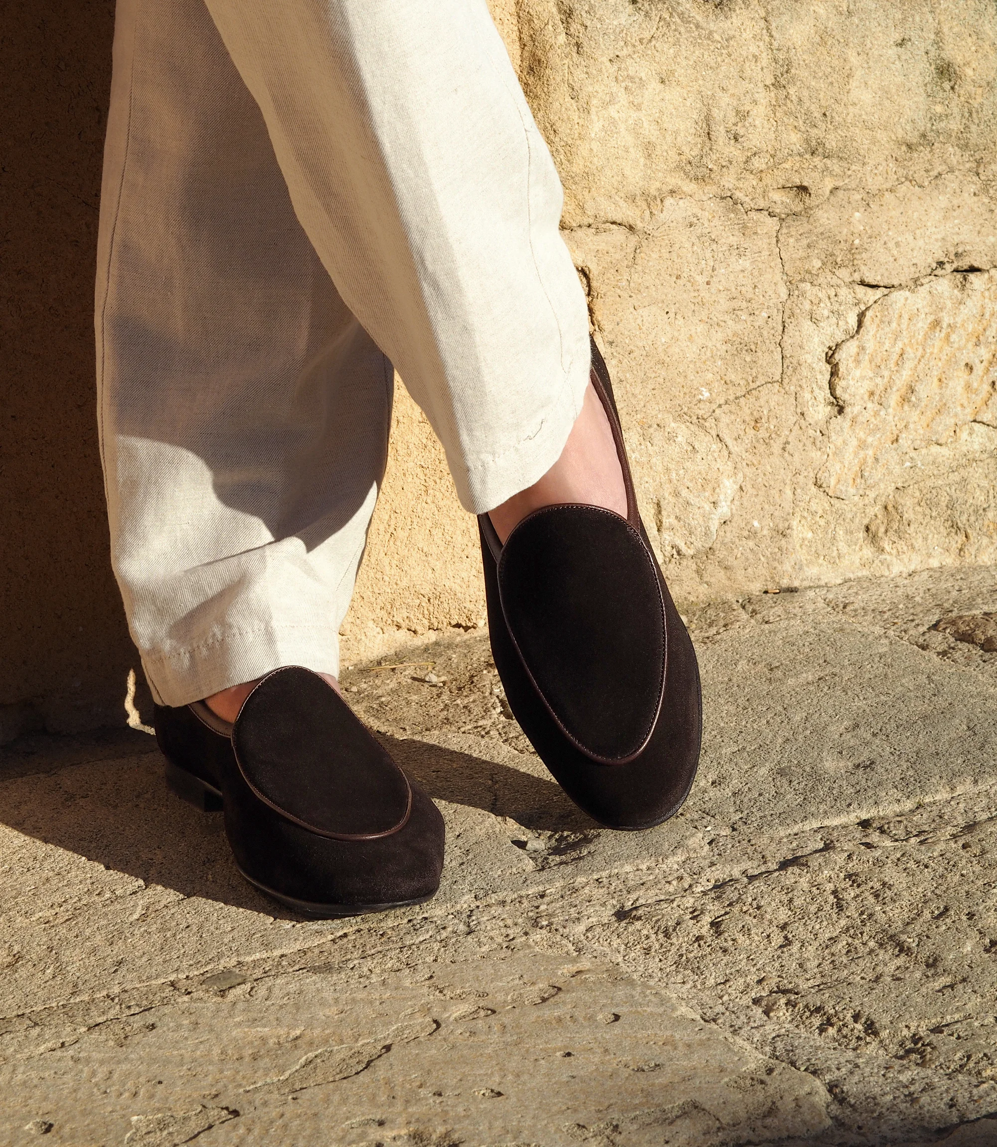 A man standing outside on holiday wearing a navy shirt and white linen trousers wearing his Loake Belgrave Loafers in dark brown suede.